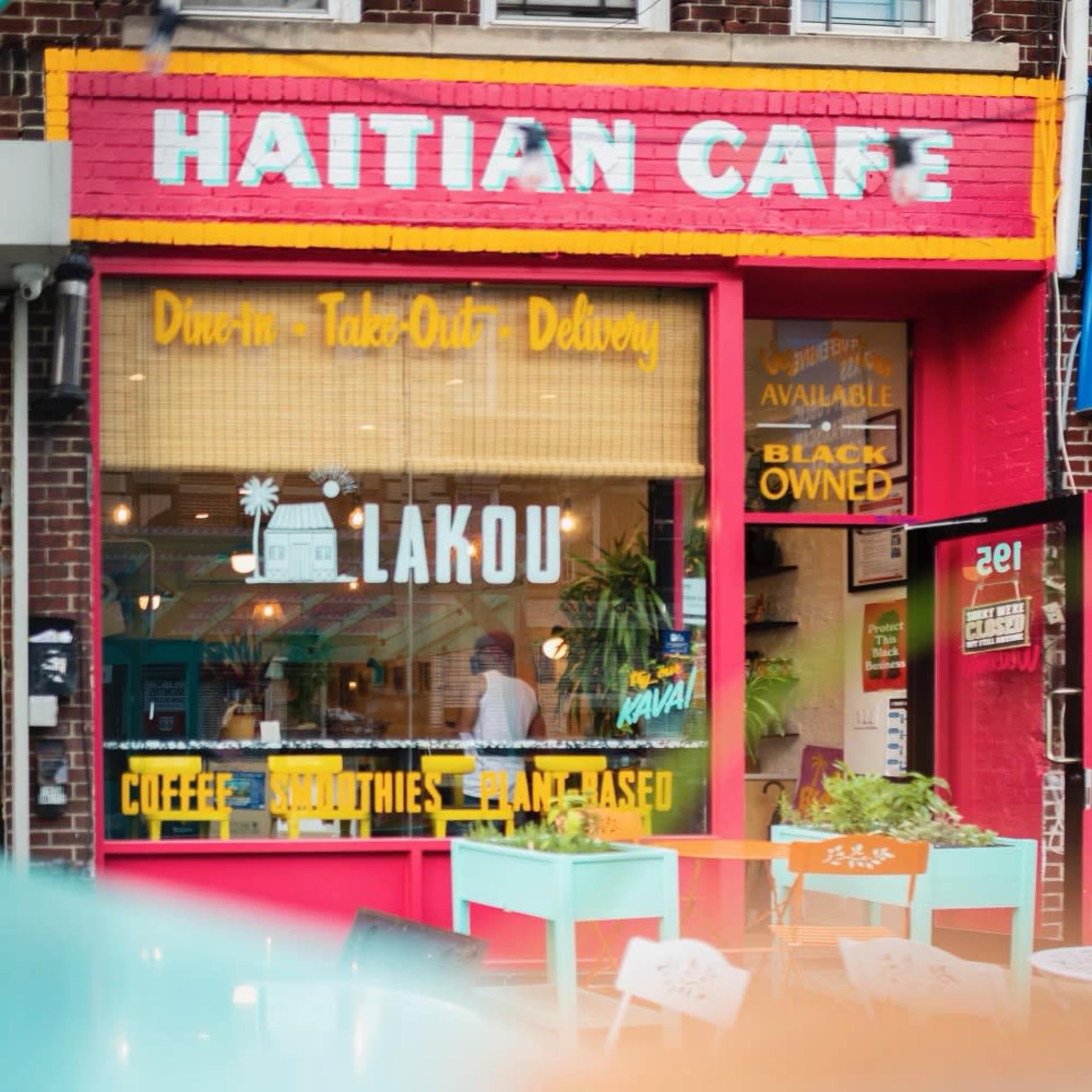 A colorful café with the sign "Haitian Café" features a large window displaying the name "Lakou" and offers coffee, smoothies, and plant-based options.