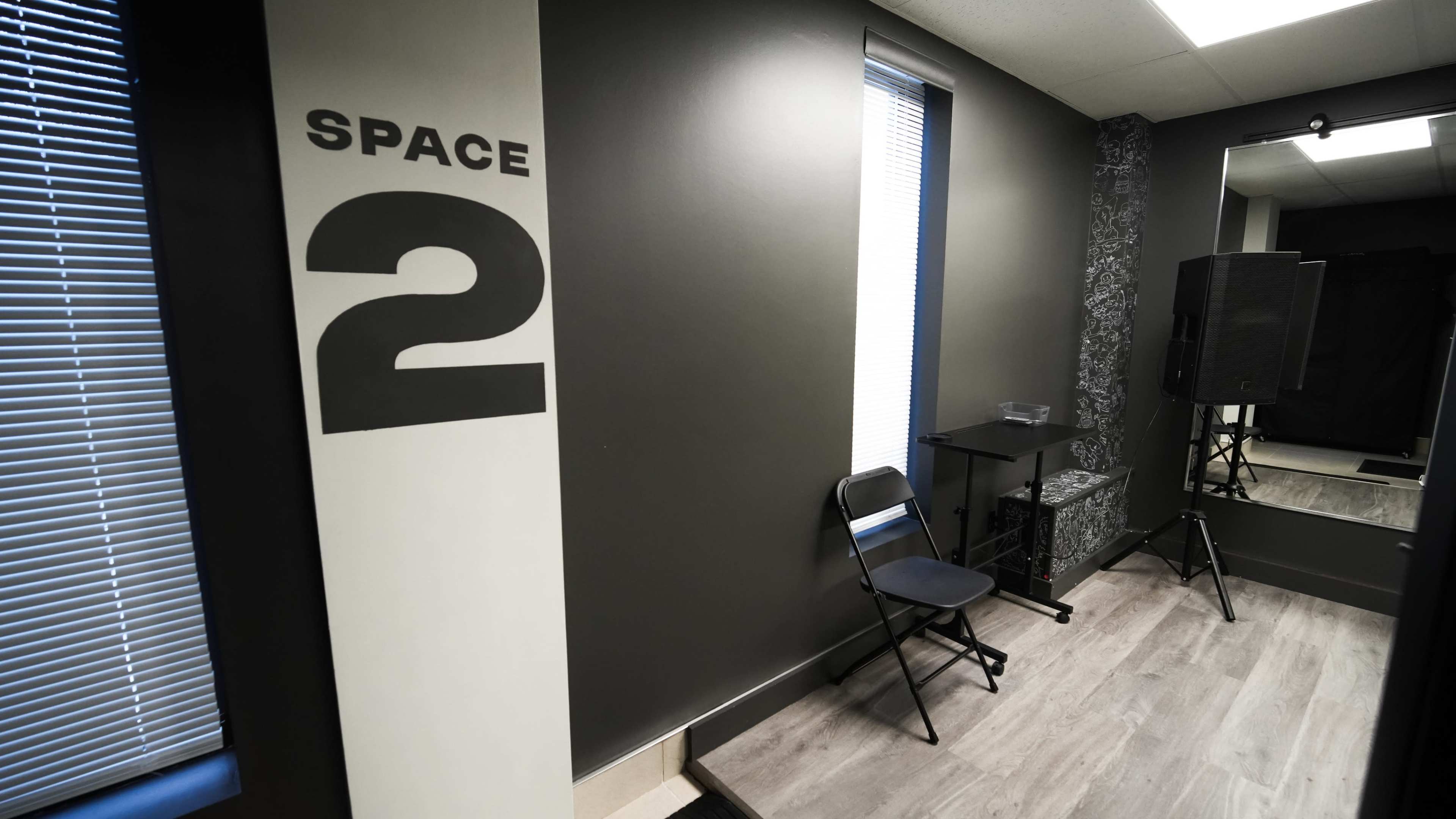 The image shows a room labeled "SPACE 2" with a black folding chair, a small table, and a mirror, alongside sound equipment against a dark wall.