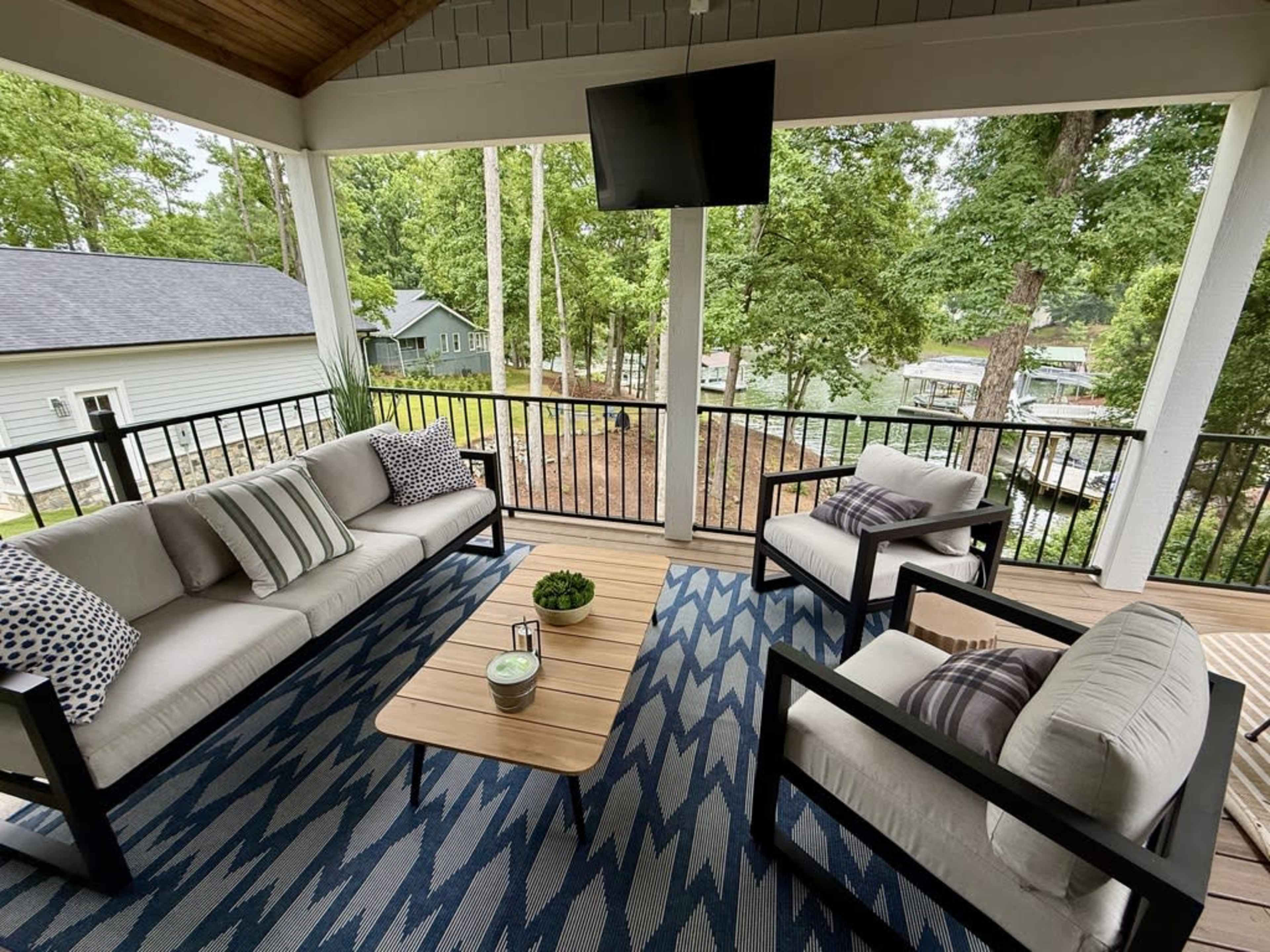 The image shows a covered porch with seating arrangements including a sofa and two chairs, a coffee table, and a large television mounted on the wall, surrounded by greenery and a view of a lake.