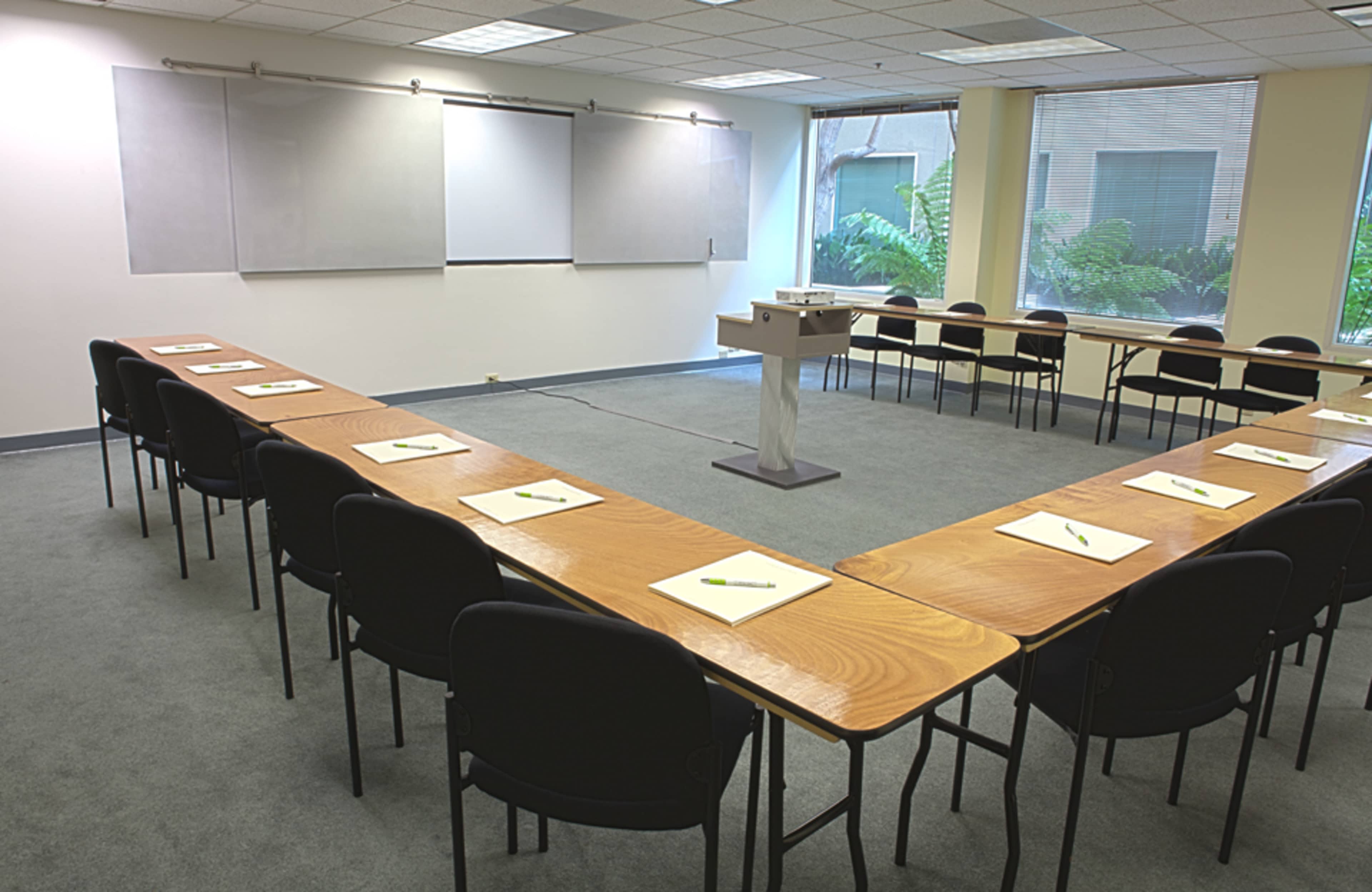 A conference room features a U-shaped arrangement of tables surrounded by chairs, with a podium at one end and whiteboards on the walls.