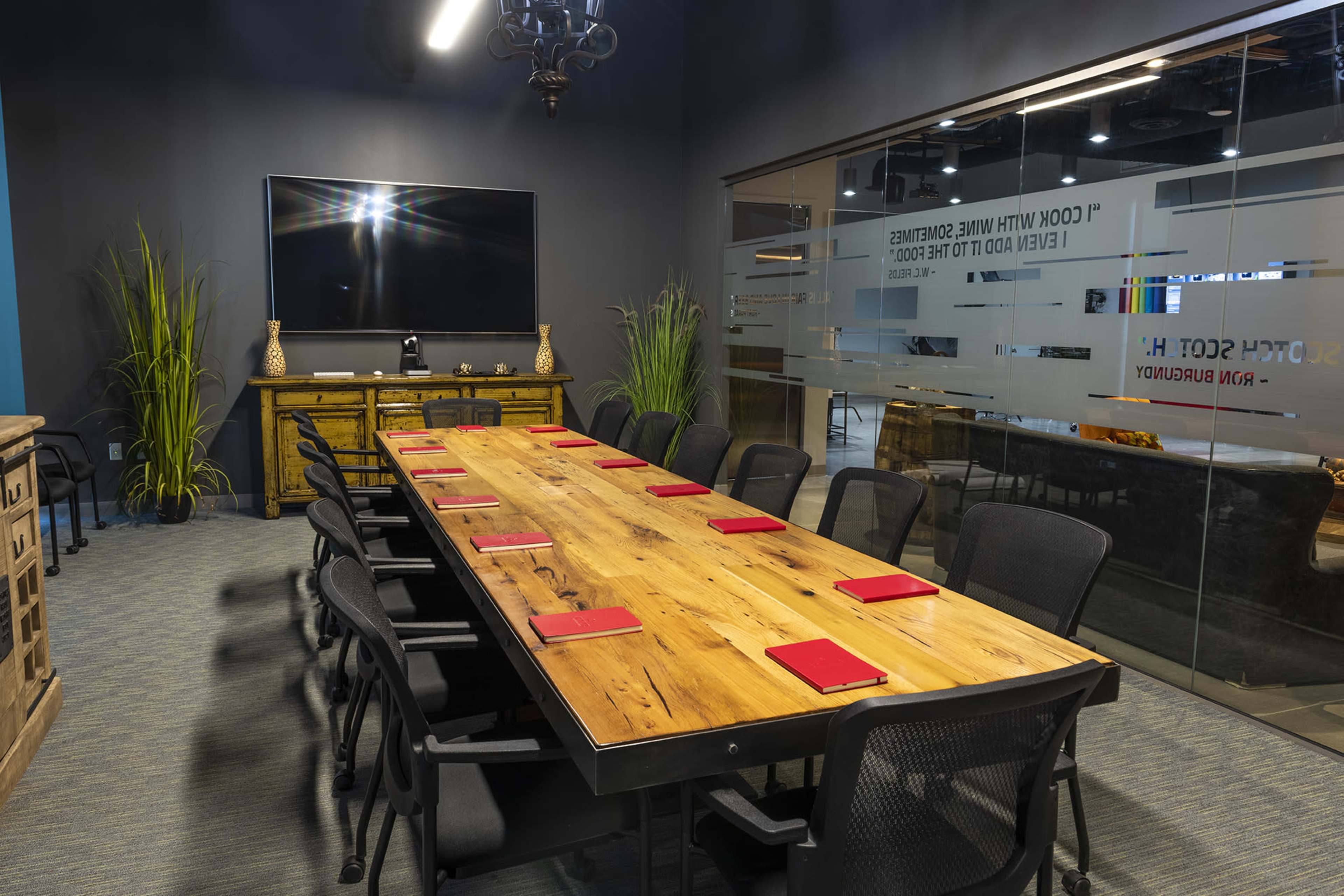 A long wooden conference table with red notepads is surrounded by black chairs in a modern meeting room featuring large windows and a wall-mounted television.