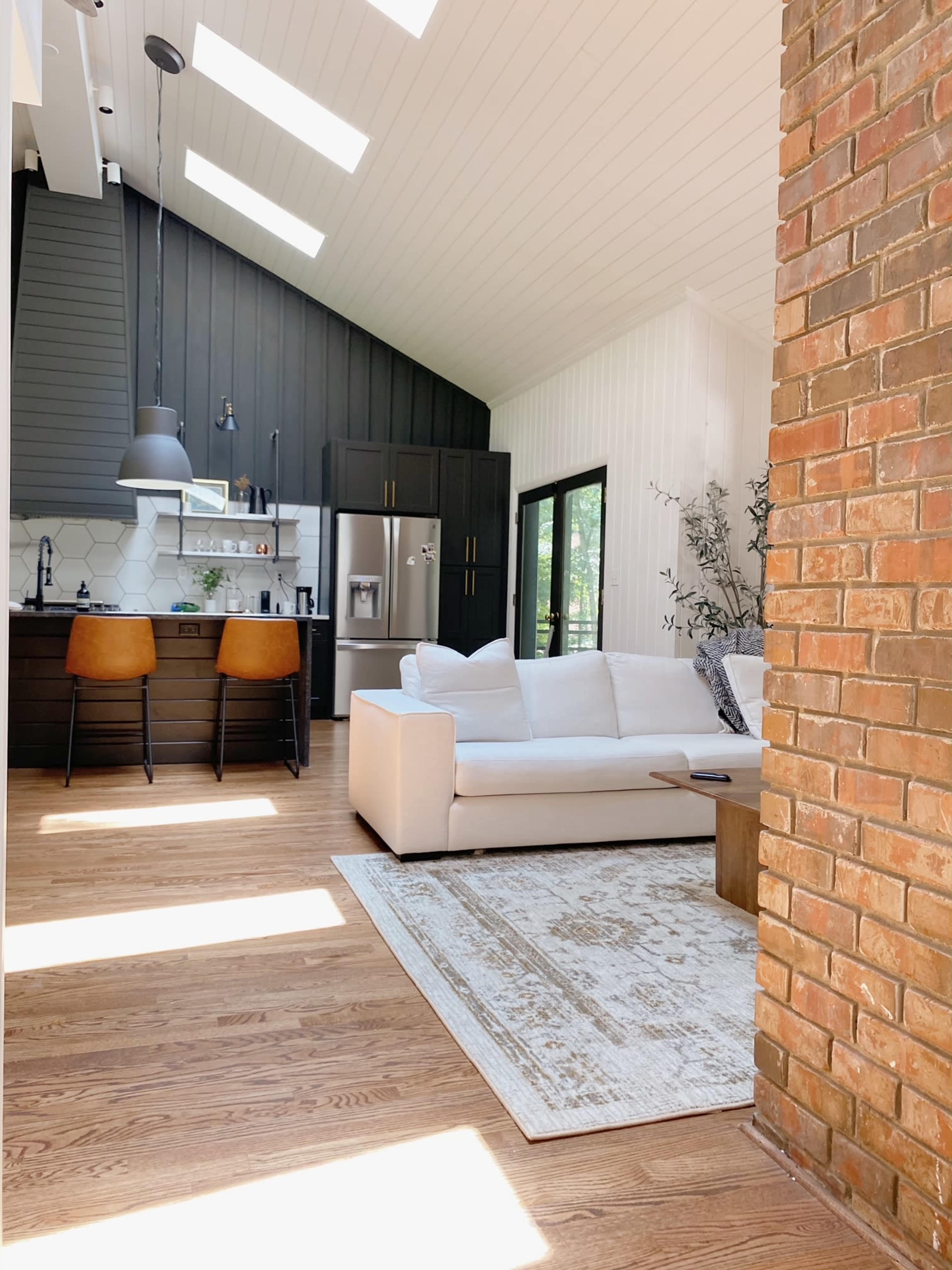 A modern living space with a light-colored sofa, a wooden table, and a kitchen area featuring dark cabinets and large windows for natural light.