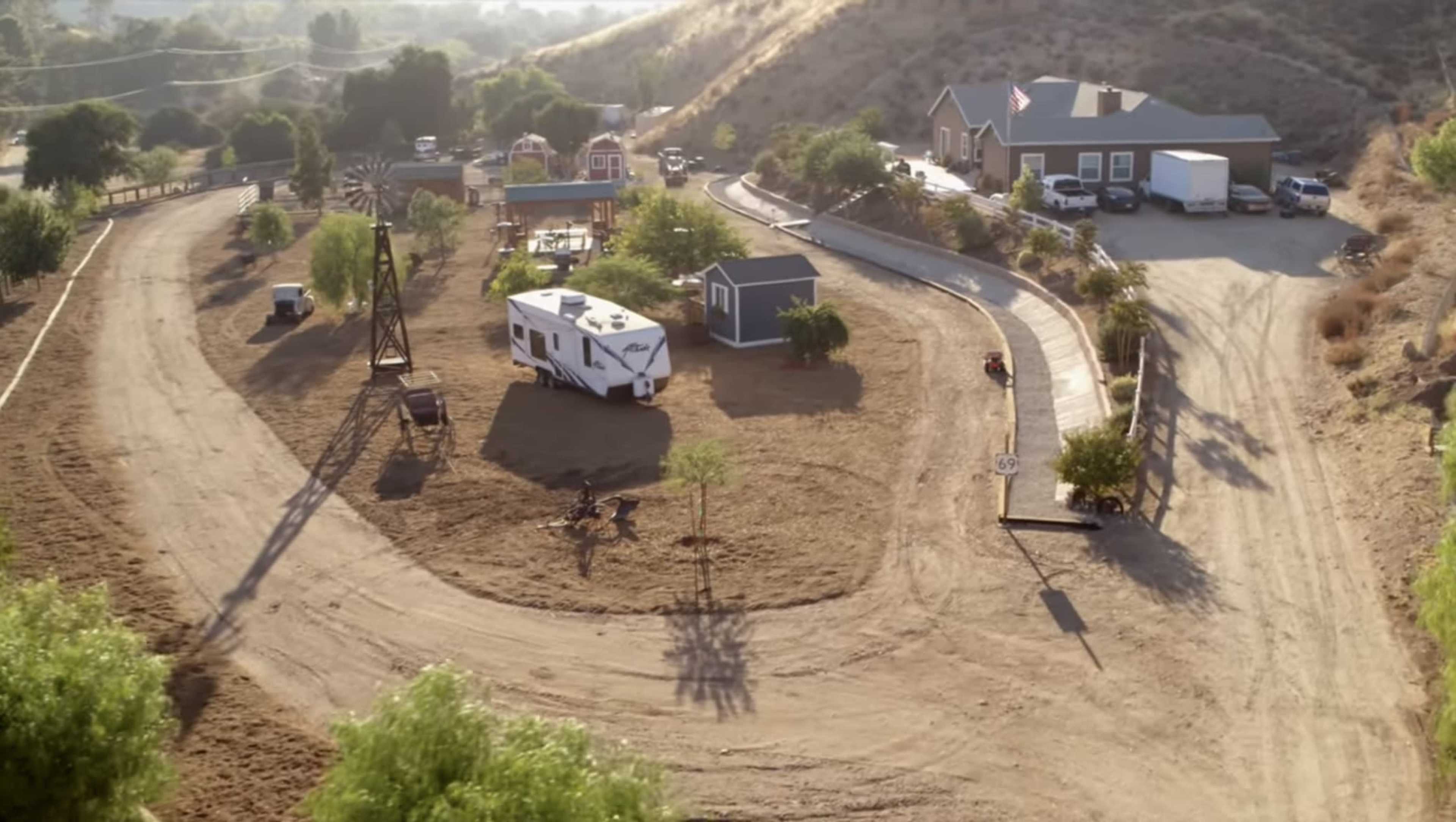 The image shows a rural property with a winding dirt driveway, a recreational vehicle parked among several trees, and a small house with additional structures in a hilly landscape.