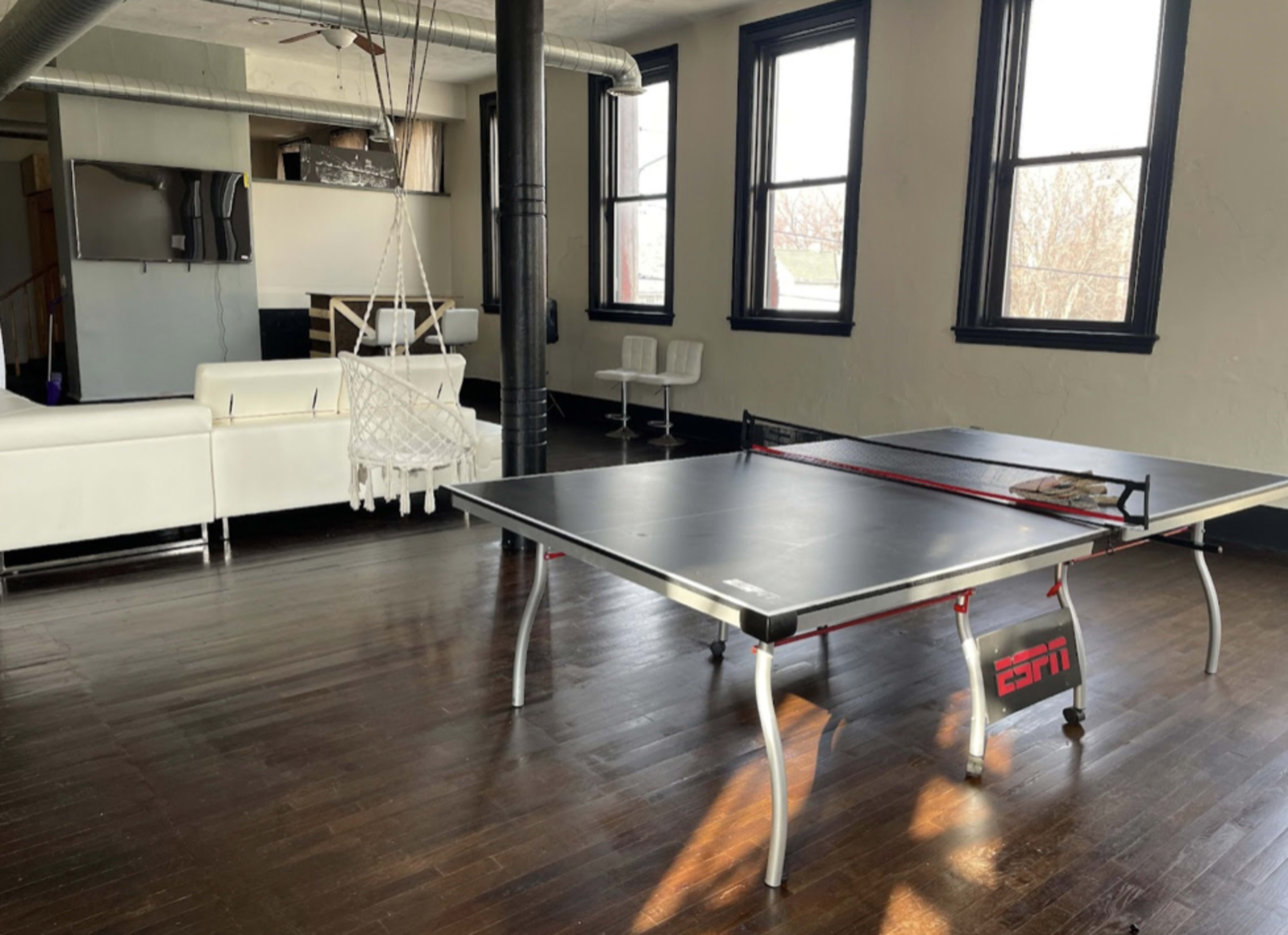 A spacious room with a ping pong table in the center, surrounded by large windows and modern furniture.