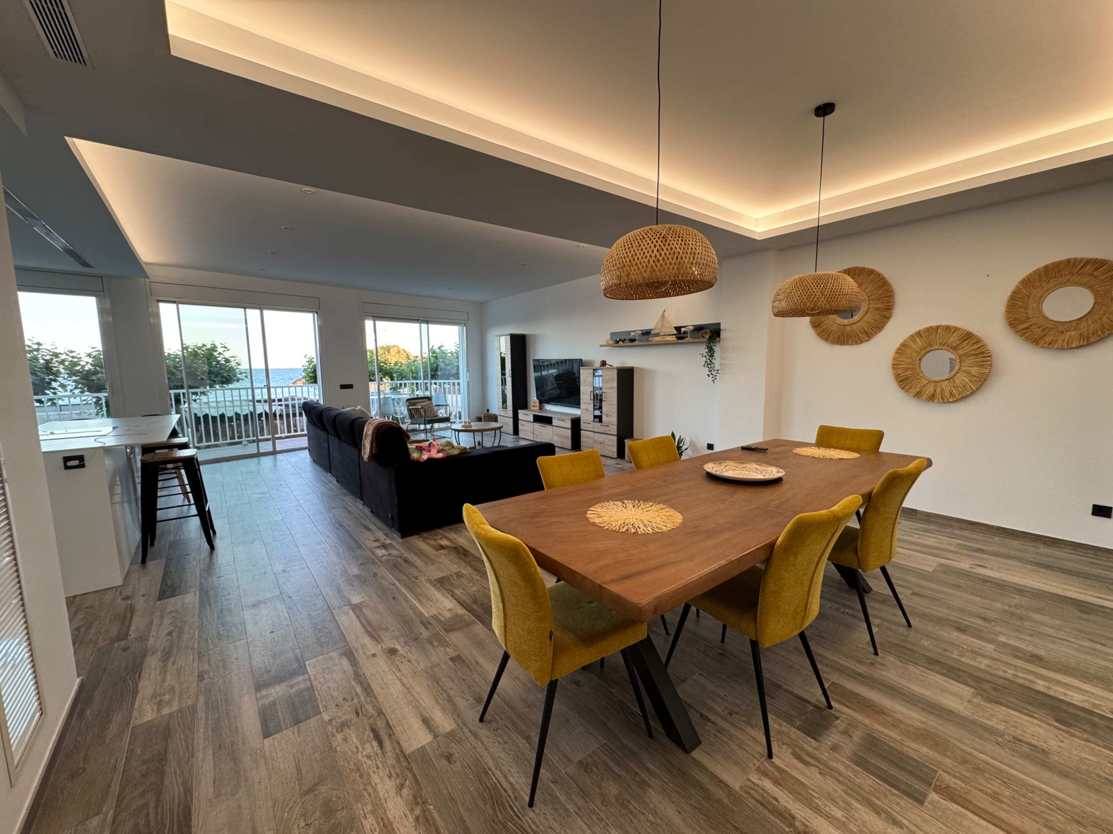 The image shows a modern living and dining area with a wooden table, yellow chairs, a dark sofa, a television, and large windows that overlook a scenic view.