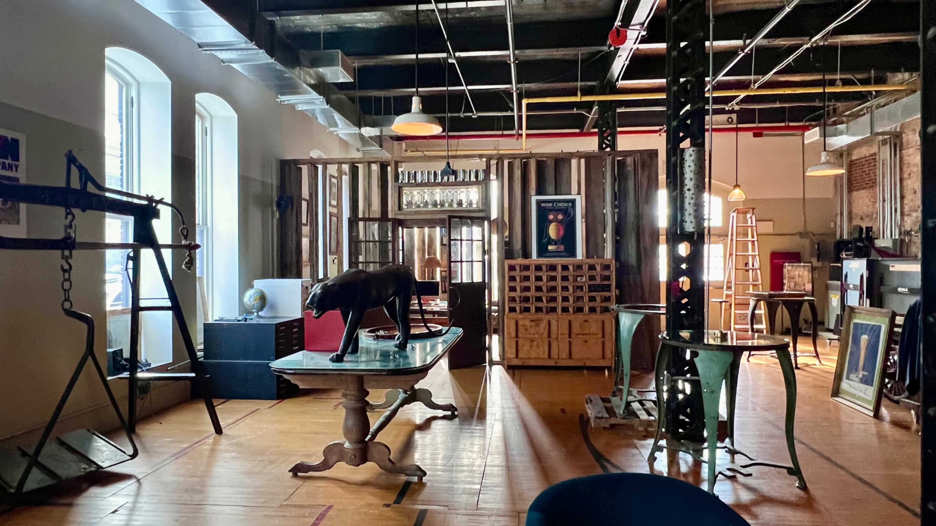 The image shows an industrial-style interior space with wooden flooring, large windows, and various pieces of furniture, including a table, a sculpture of a dog, and shelves stacked with boxes.
