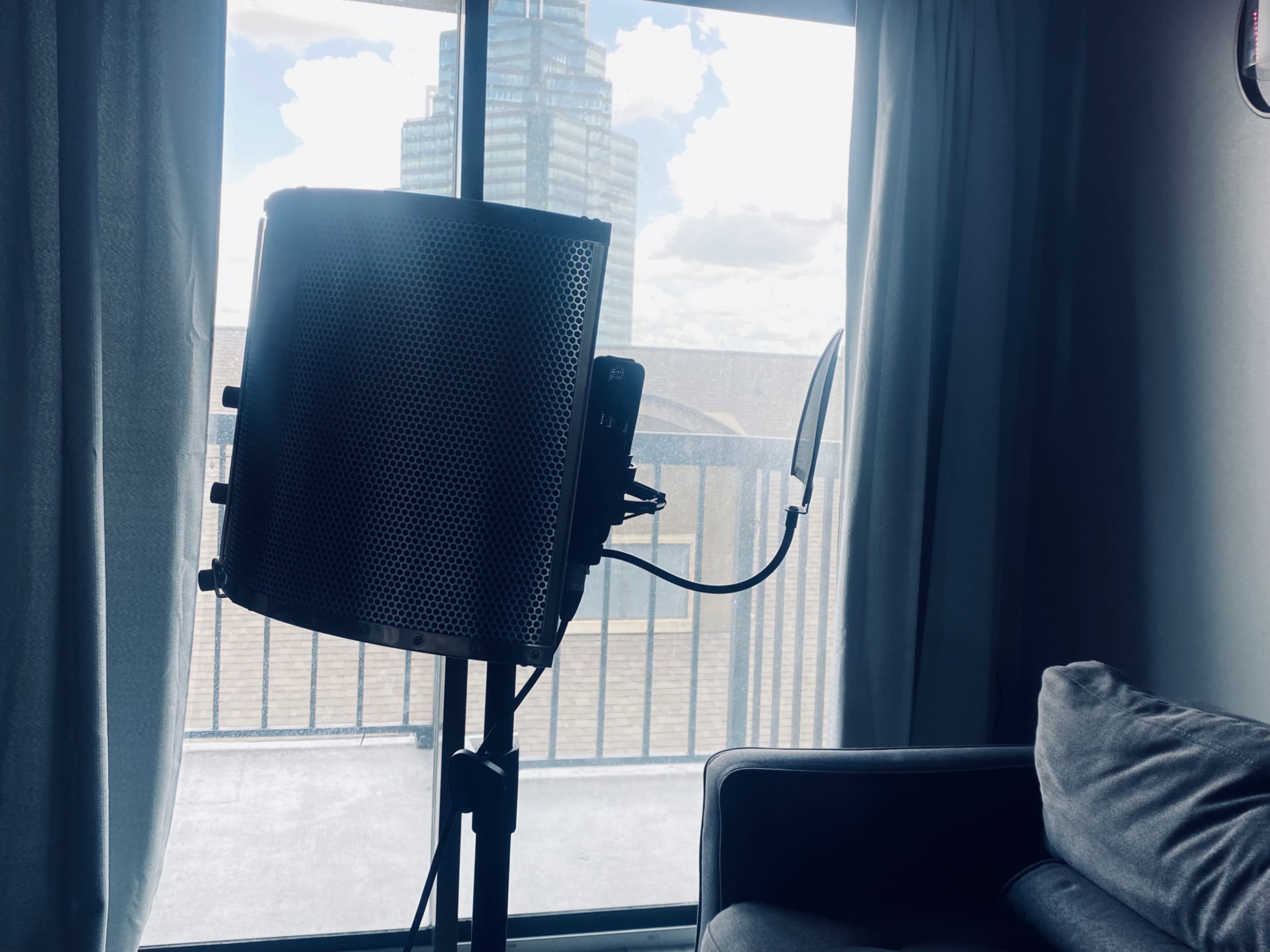 A microphone stand with a pop filter is set up near a window overlooking a building and a partly cloudy sky.
