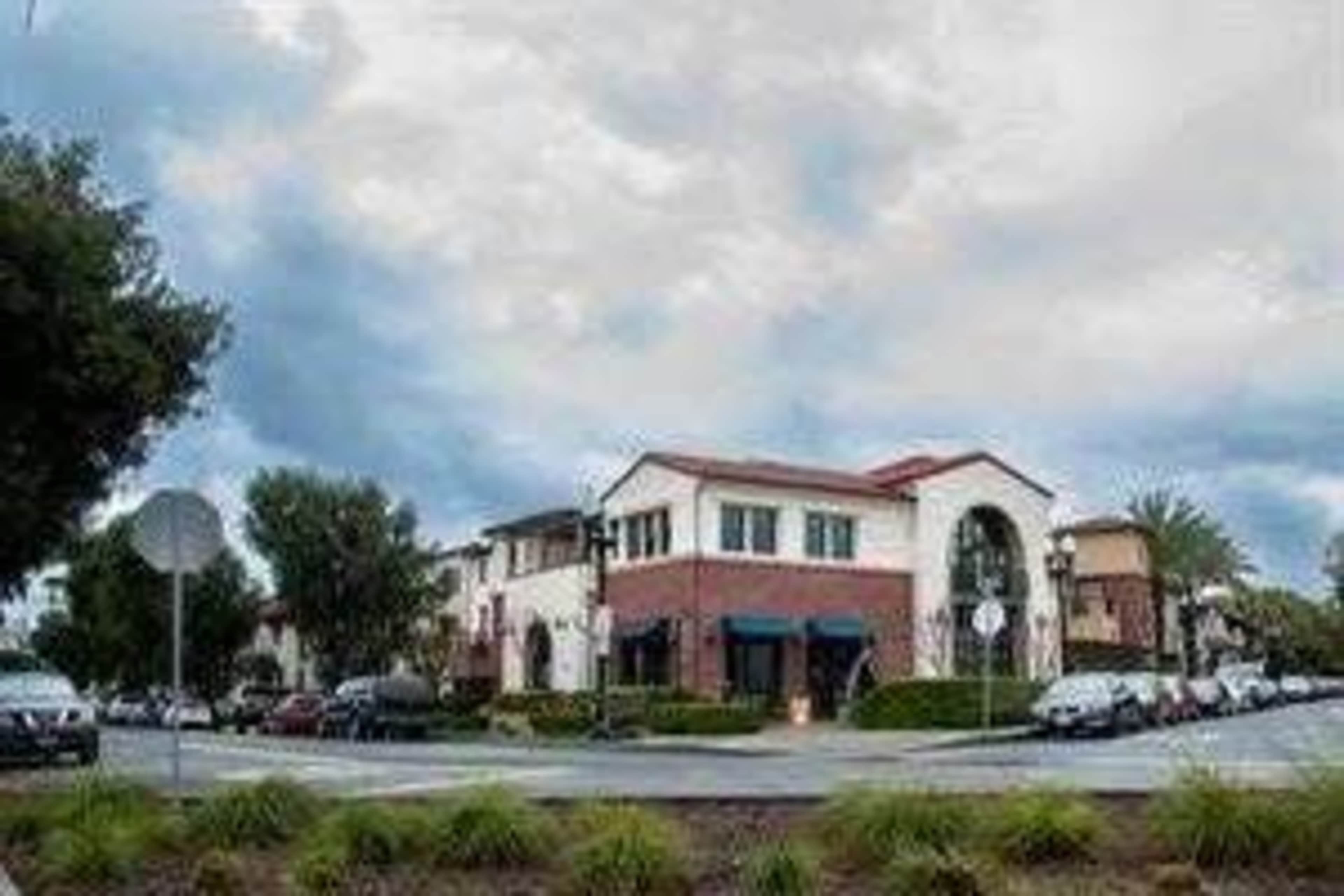 The image shows a street scene featuring a mixed-use building at a roundabout, surrounded by parked cars and palm trees under a cloudy sky.