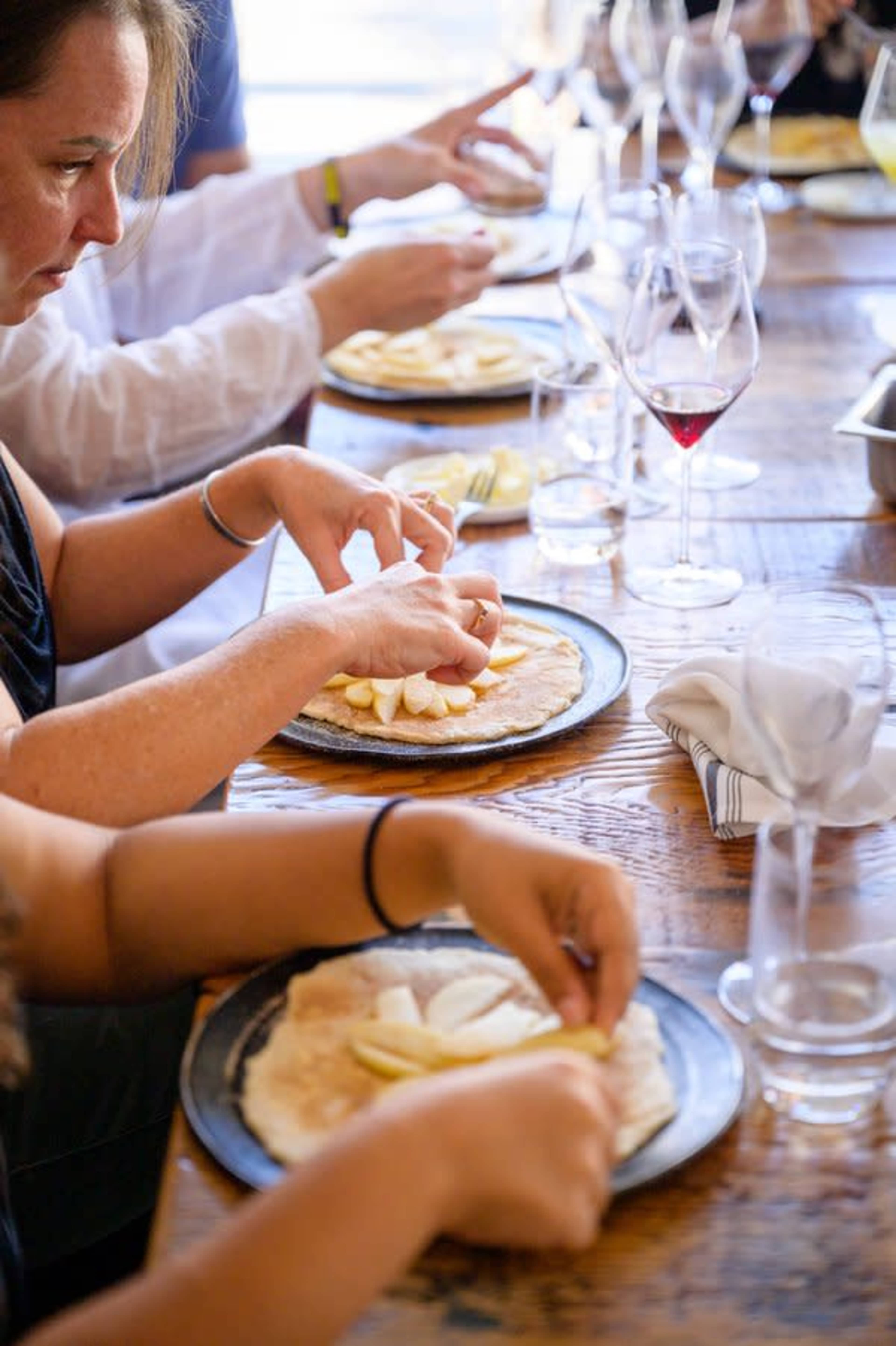 People are preparing dessert by arranging slices of fruit on flatbread at a dining table.