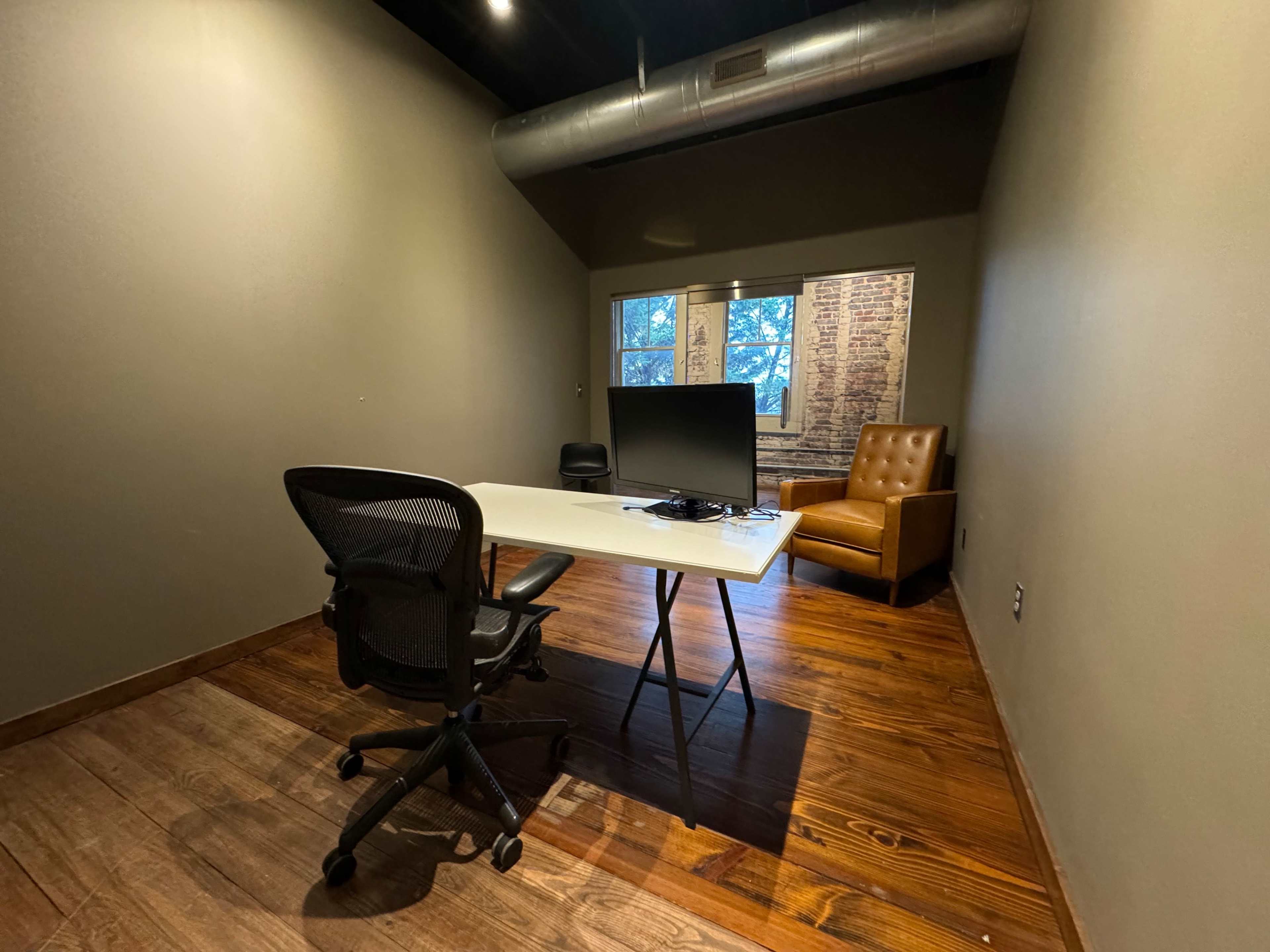 The image shows a small office space with a desk, a rolling chair, a flat-screen monitor, a brown armchair, and two windows allowing natural light to enter.