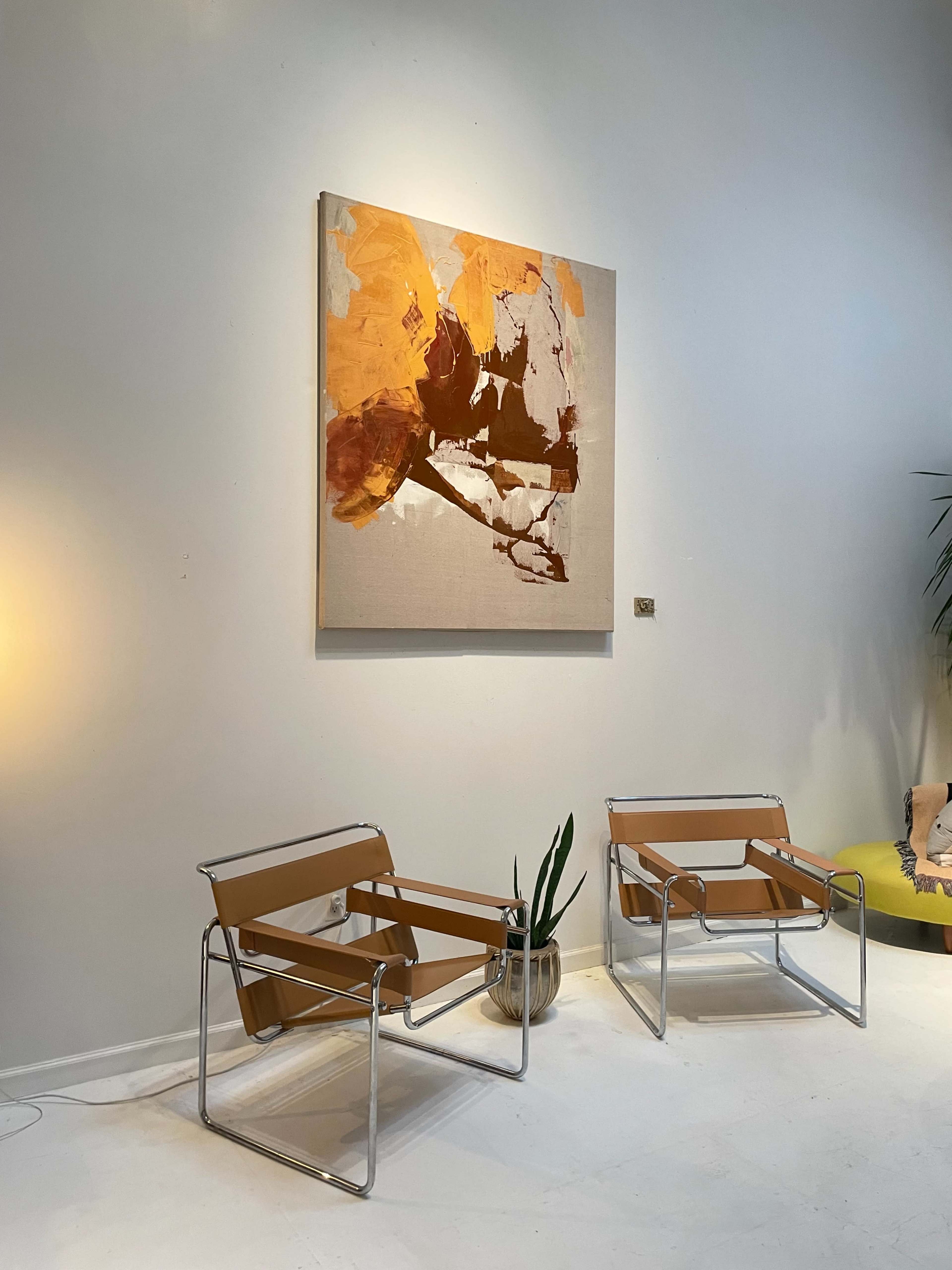 Two metal-framed chairs with tan fabric sit opposite a large abstract painting in warm tones on a pale wall, accompanied by a potted plant nearby.