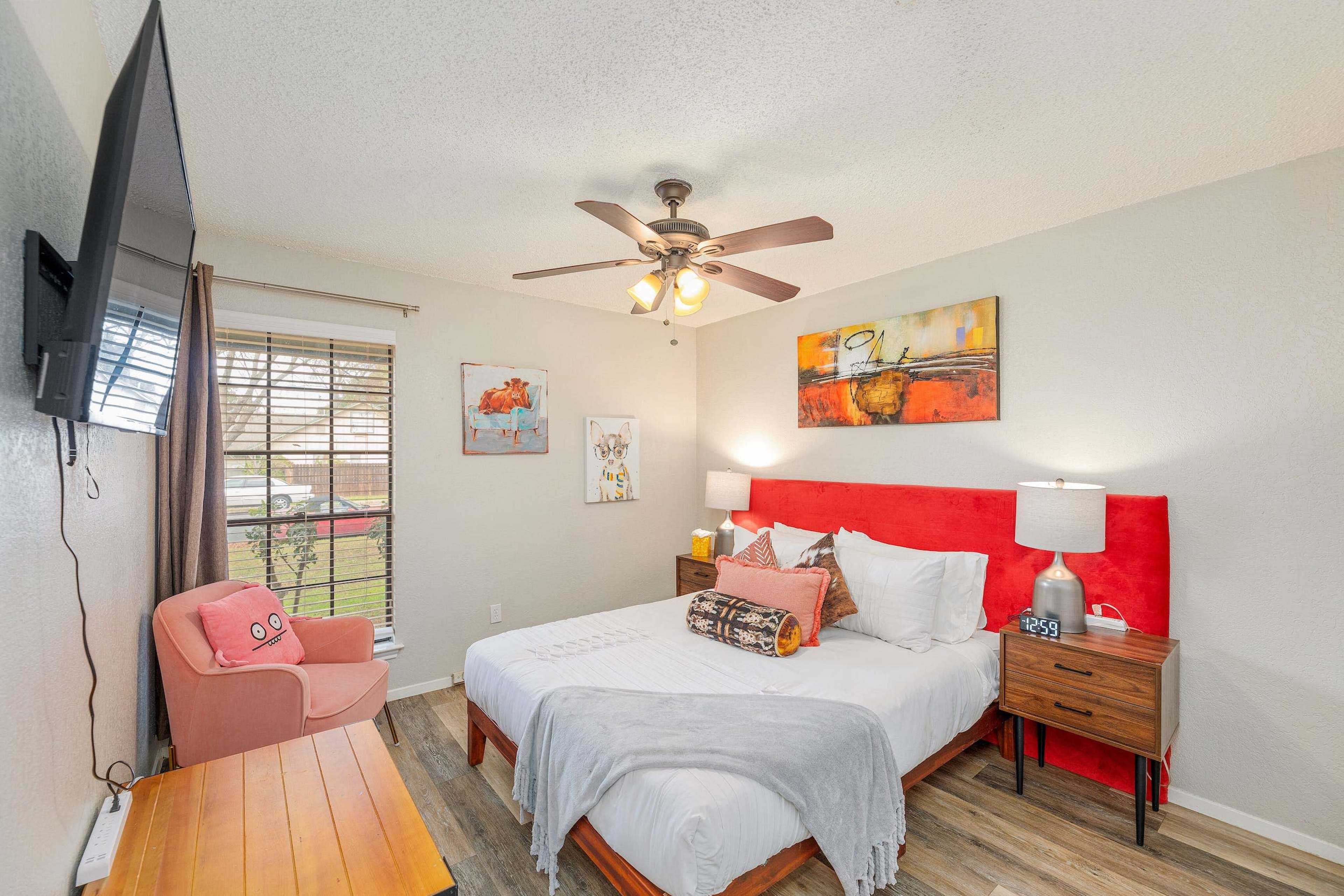 The image shows a bright bedroom featuring a queen-size bed with a red headboard, two nightstands with lamps, a television mounted on the wall, and a window with blinds.