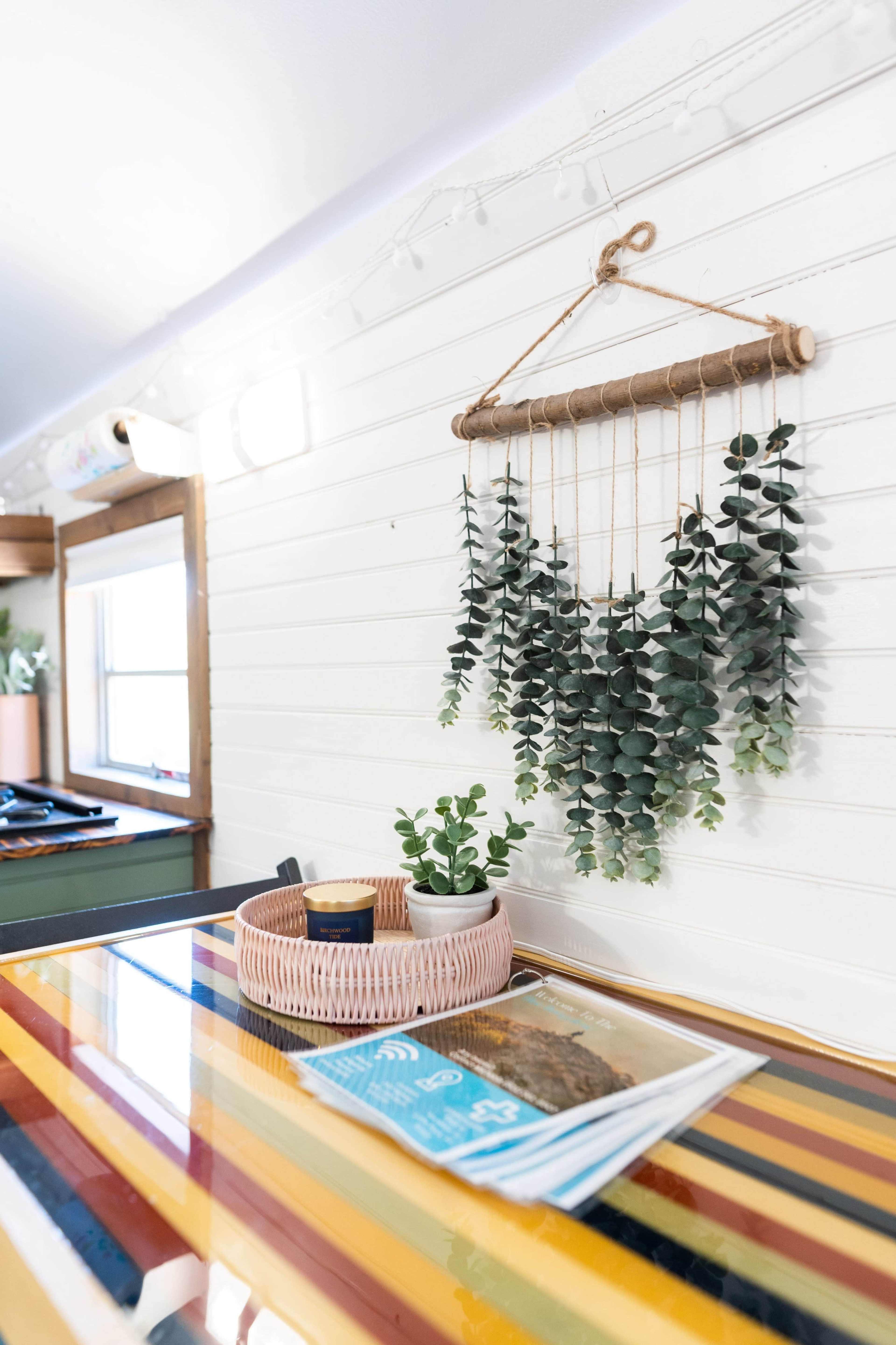 A colorful striped table displays magazines alongside a pink woven basket and hanging greenery against a white wall.