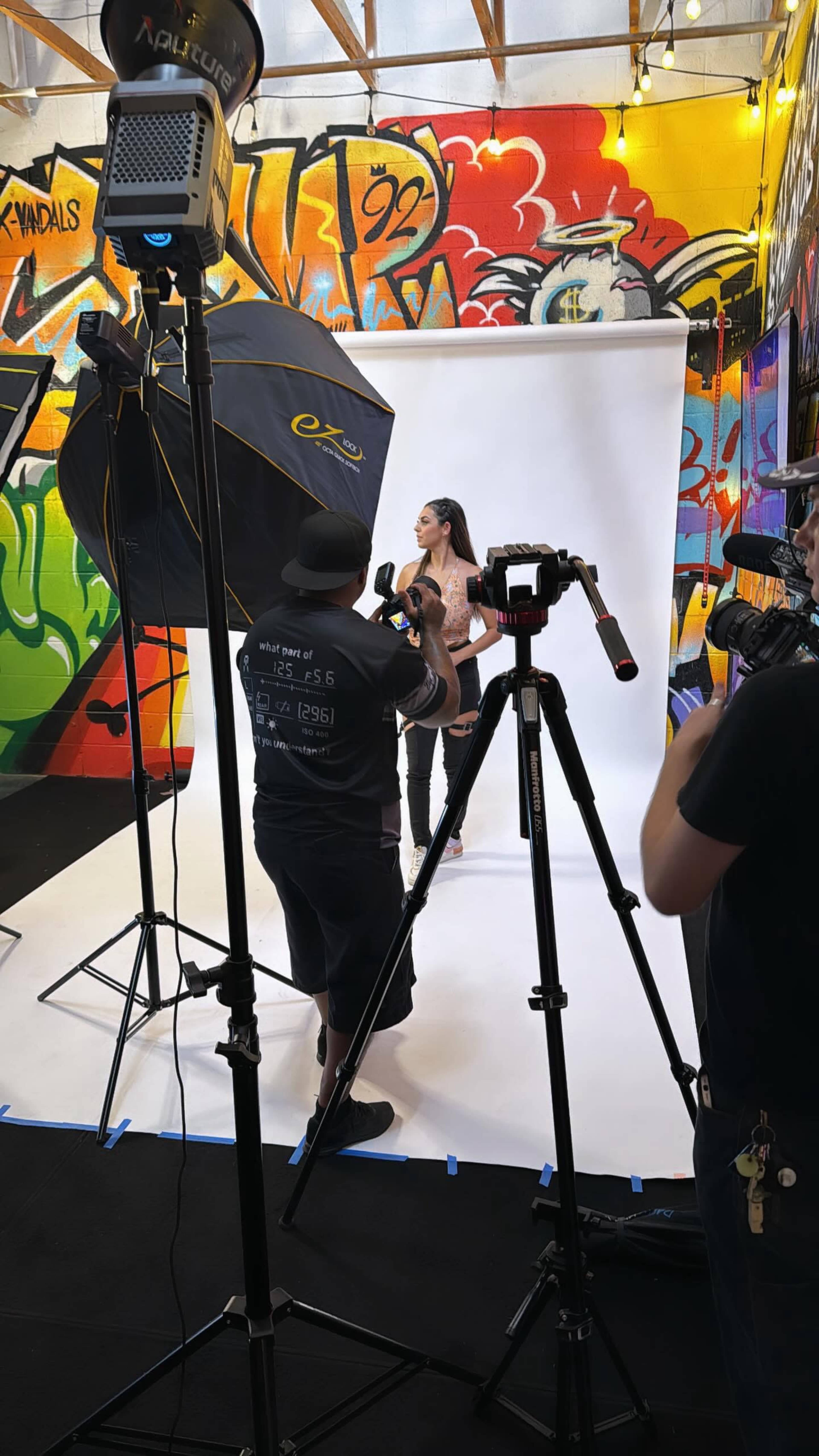A model poses in front of a white backdrop while photographers and lighting equipment surround her in a graffiti-decorated studio.