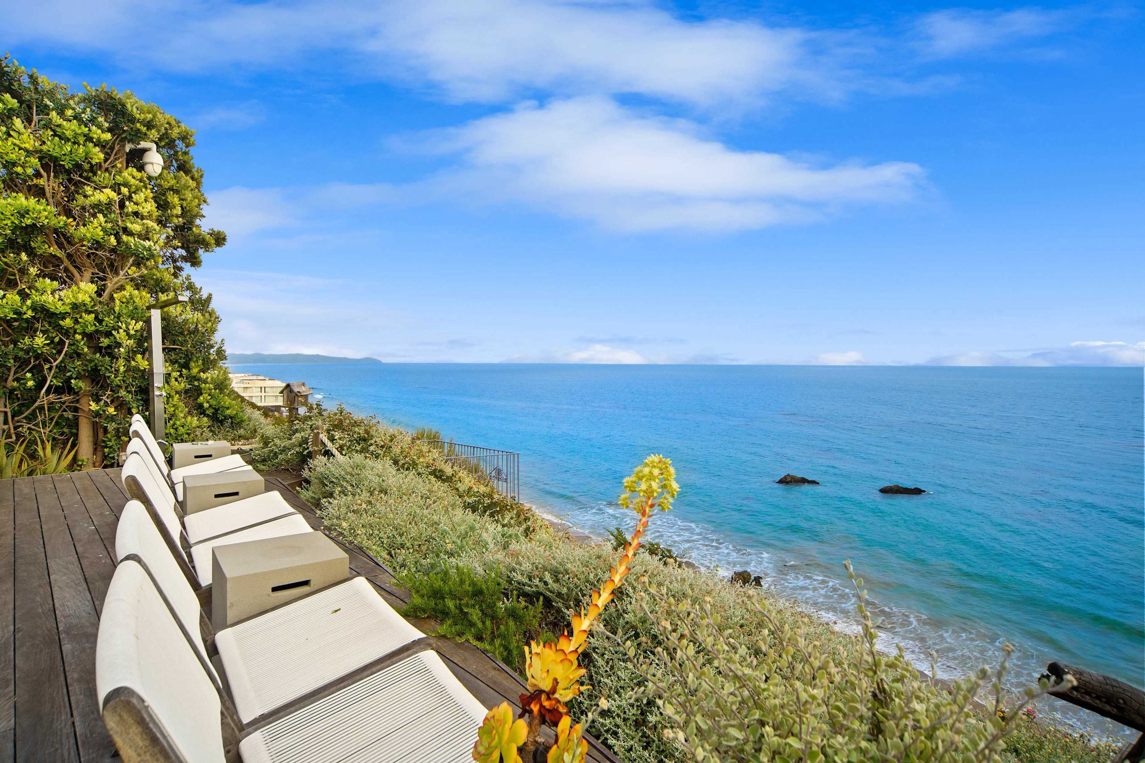 A row of white lounge chairs lines a wooden deck overlooking a calm ocean with rocky outcrops in the distance.