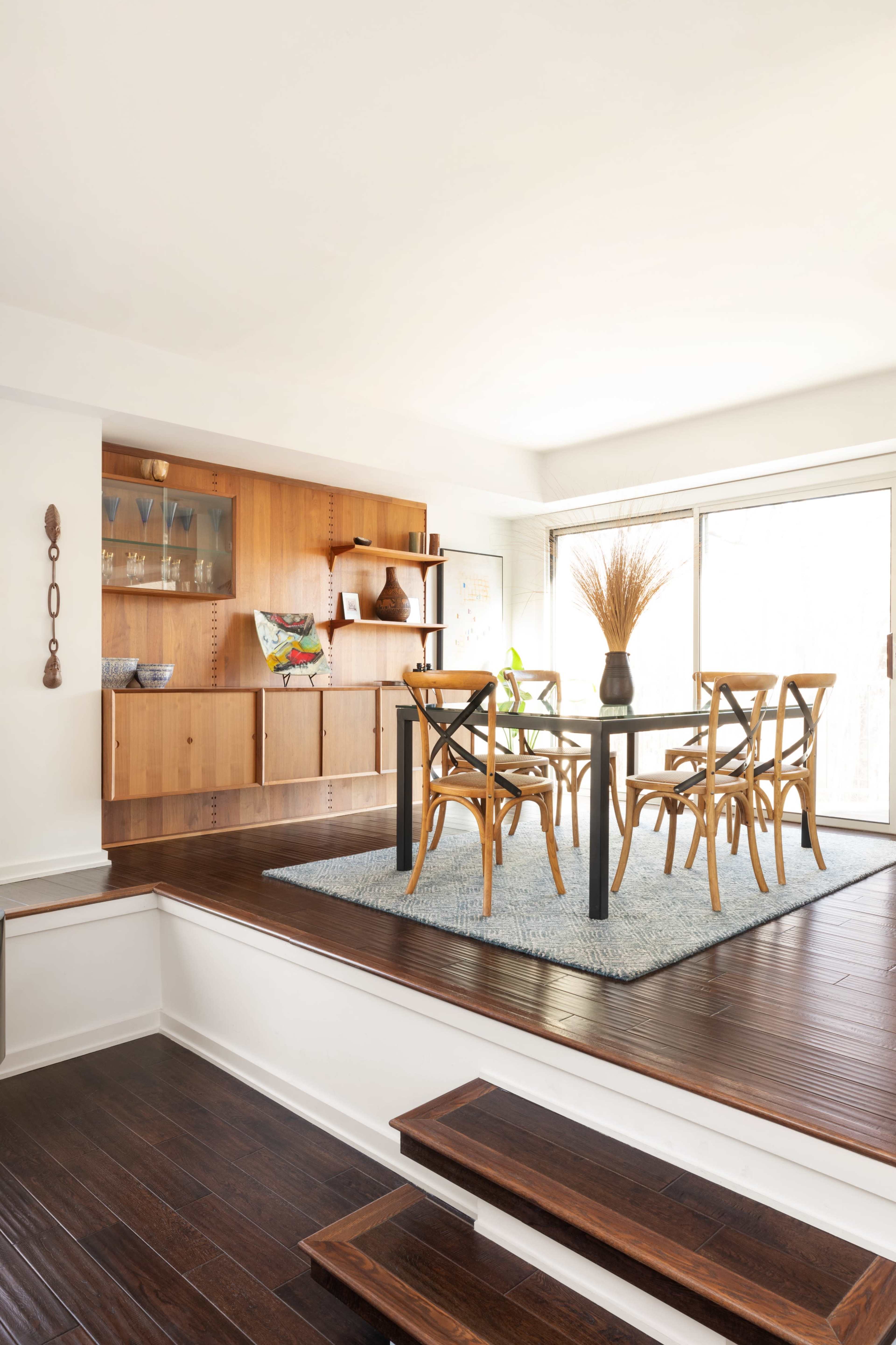 A light-filled dining area features a glass table surrounded by wooden chairs, with a wooden cabinet and large windows in the background.