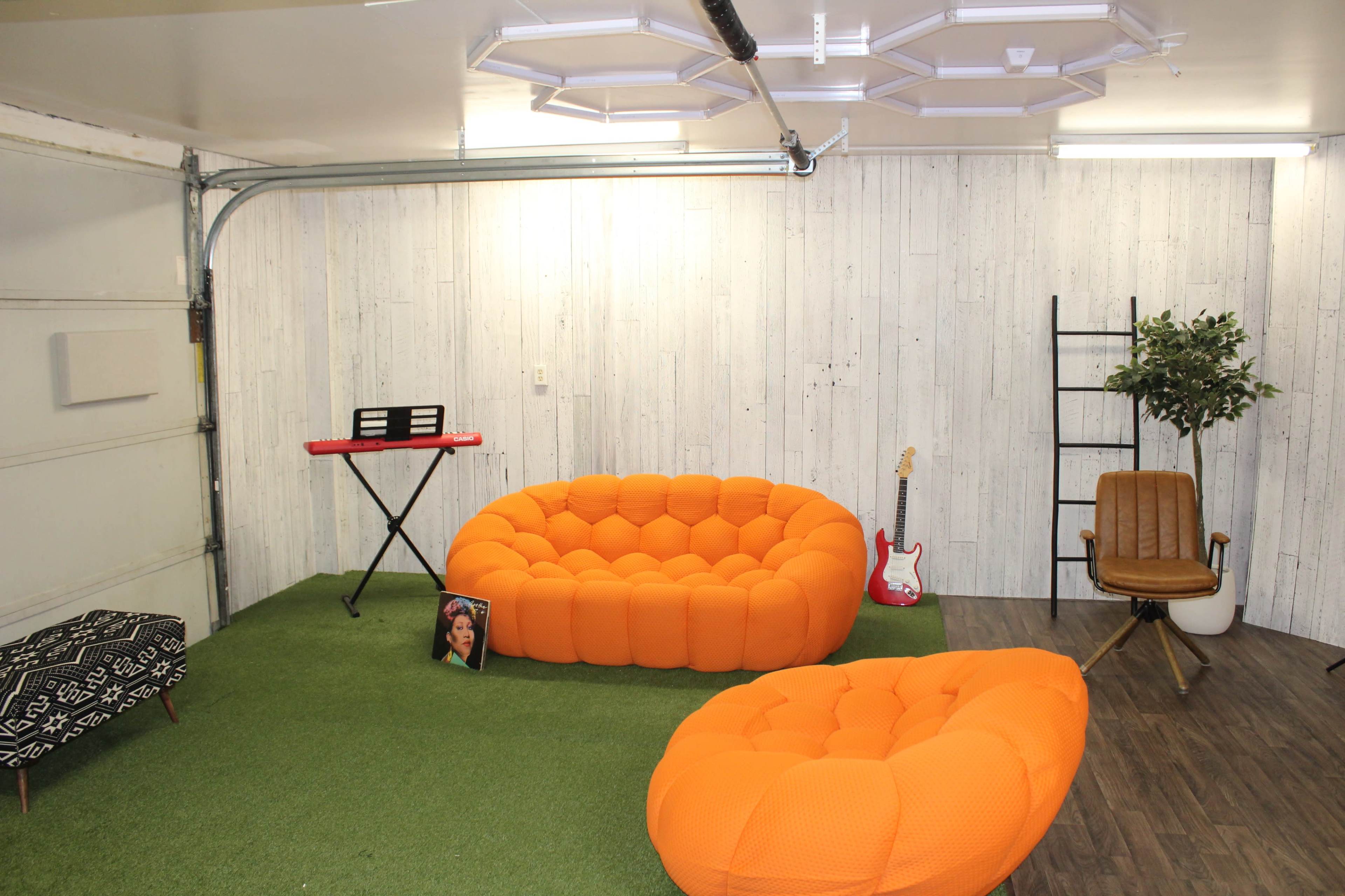 The image shows a small indoor space featuring two bright orange sofas, a keyboard, a guitar, a wooden chair, a shelf, and an artificial plant, all on a grassy floor.