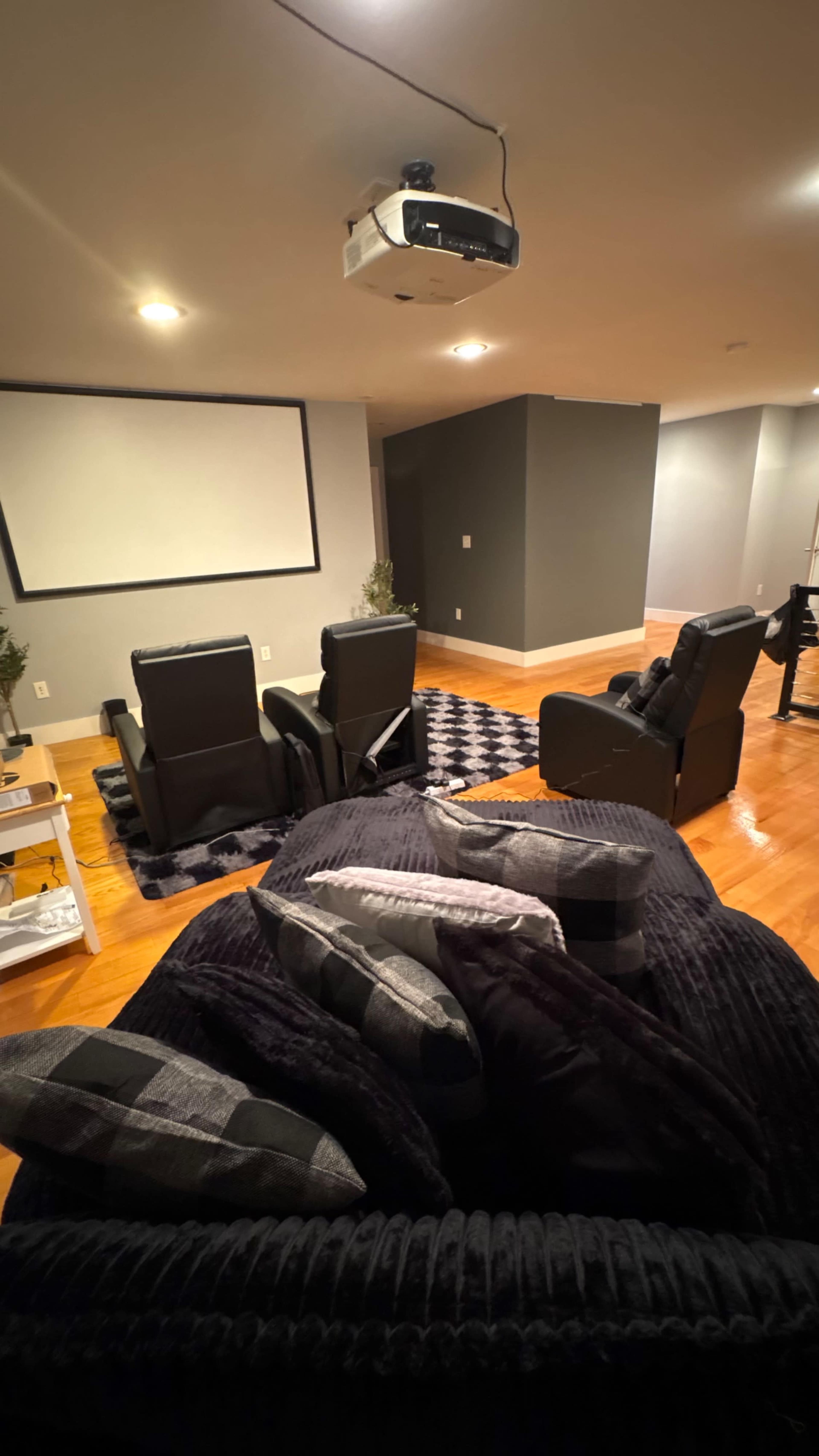 The image shows a cozy home theater with dark walls, a projector mounted on the ceiling, and two recliner chairs facing a blank projection screen.
