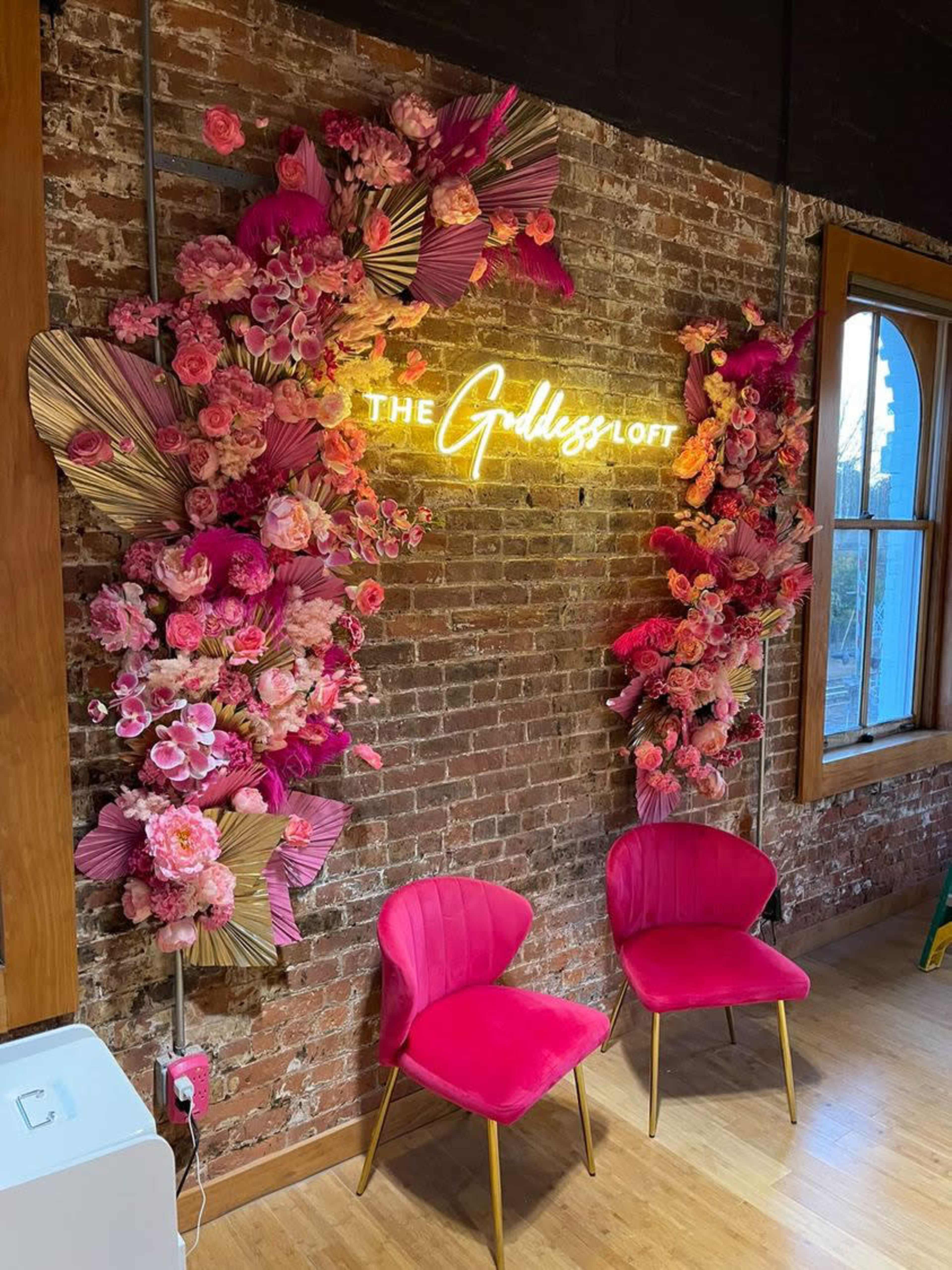 A brick wall adorned with vibrant pink floral arrangements and a neon sign that reads "The Goddess Loft," accompanied by two pink chairs.