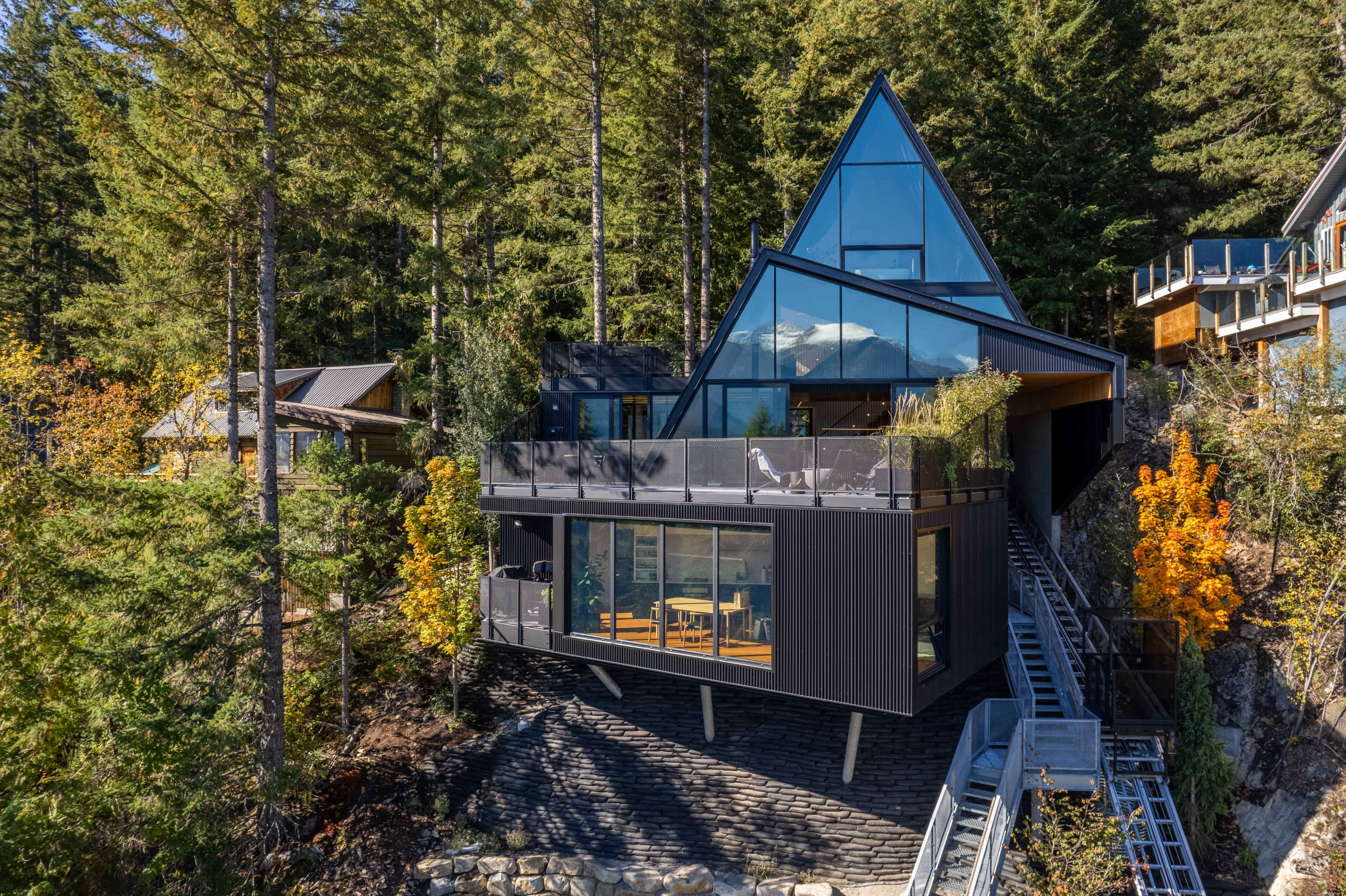 A modern triangular-roofed house with large glass windows is situated on a rocky slope surrounded by tall trees.