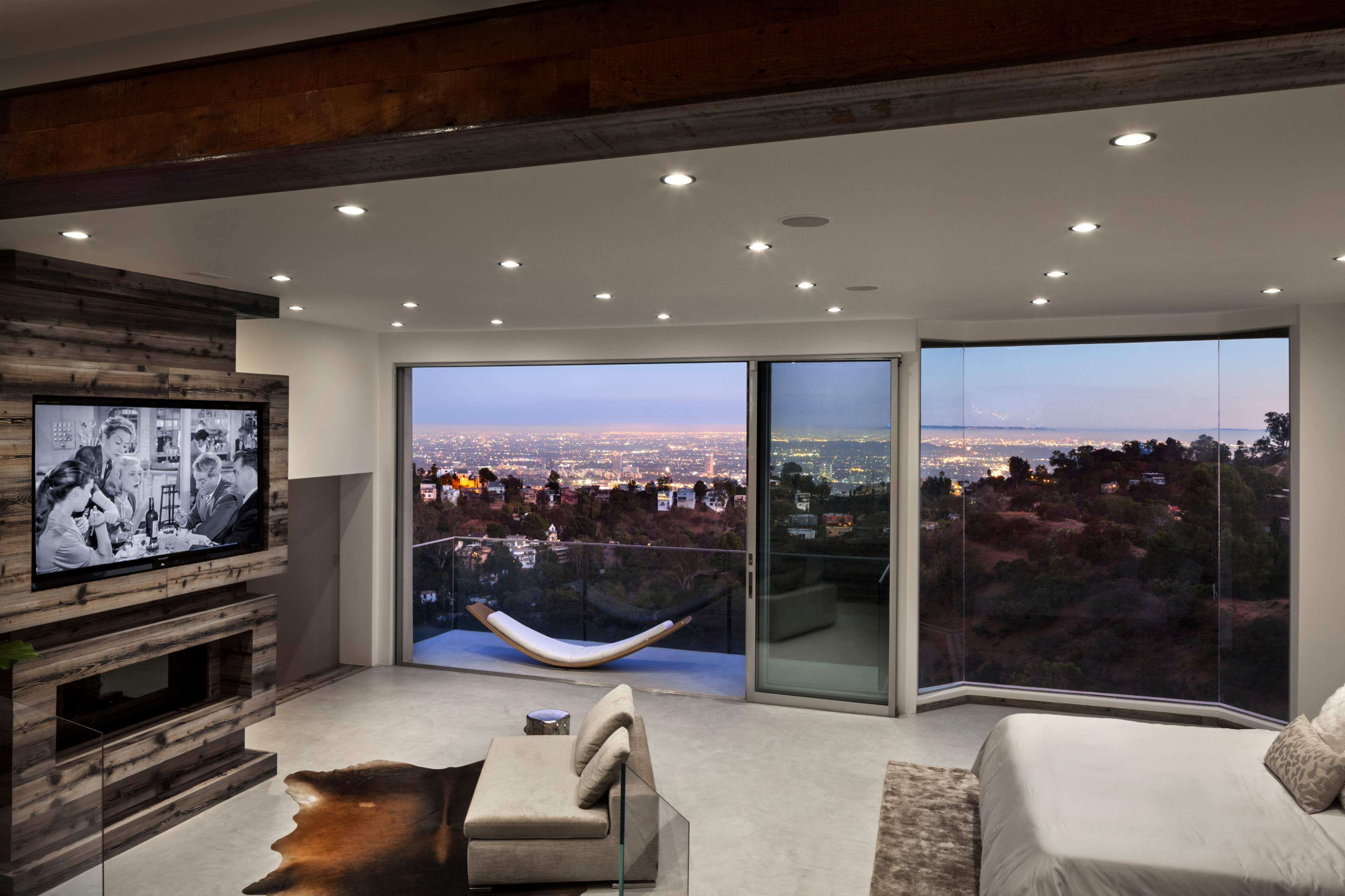 A modern bedroom features a large window offering a view of a city skyline illuminated at night, with a television mounted on the wall and a hammock suspended near the glass doors.