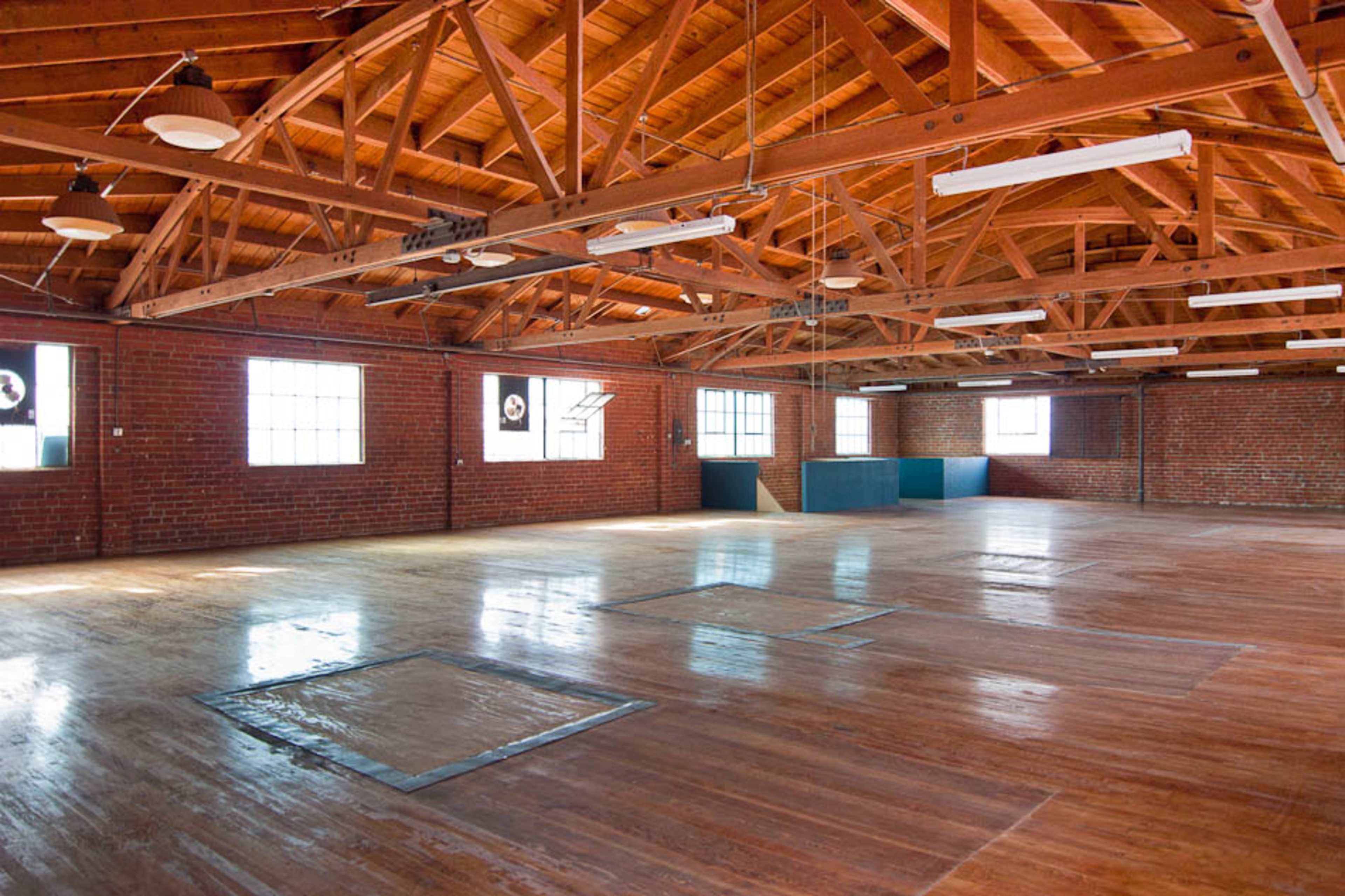 A spacious, empty warehouse interior with polished wooden floors and exposed wooden beams supporting the ceiling.