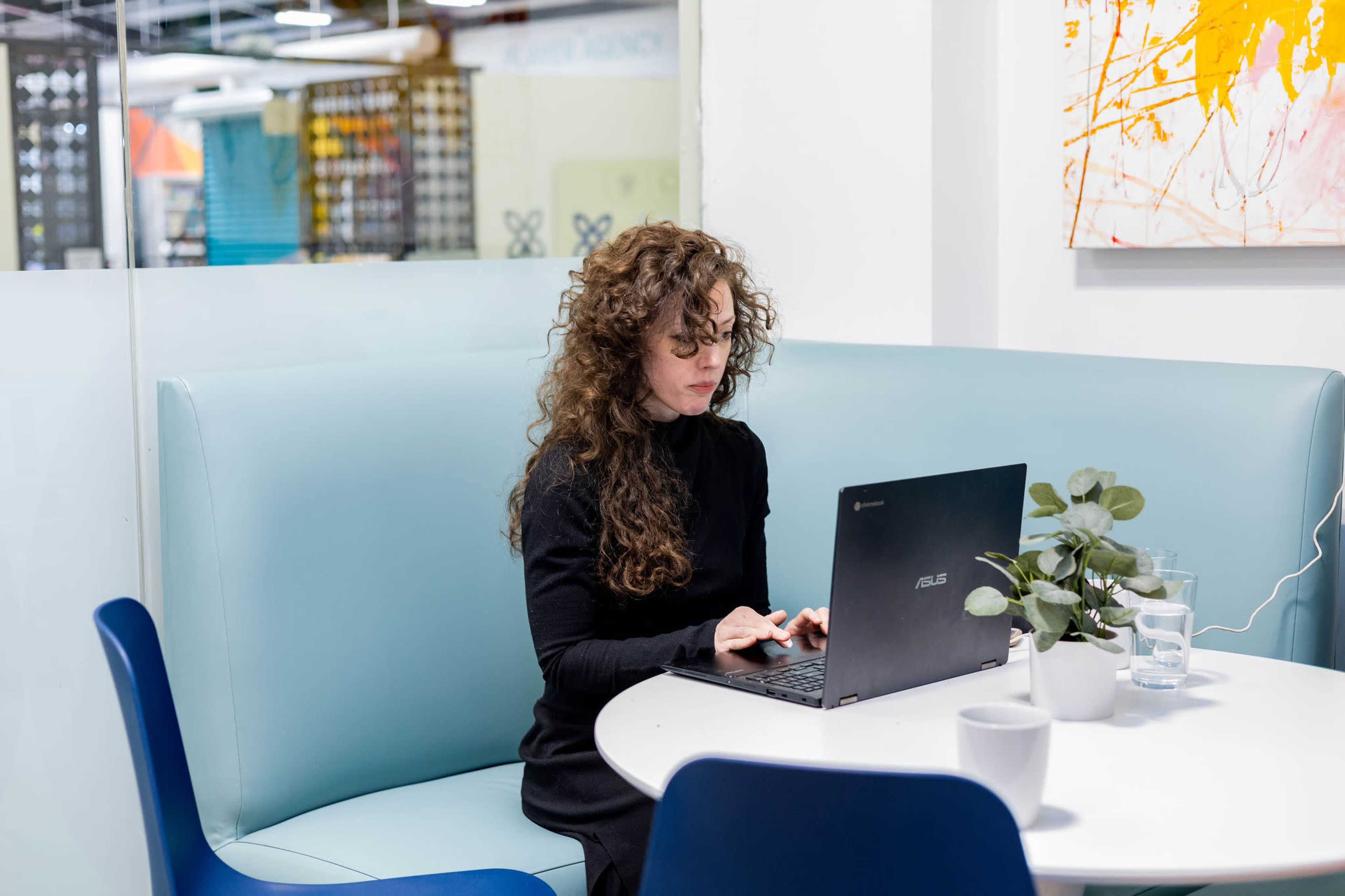 A woman with curly hair is working on a laptop at a round table in a modern office setting.