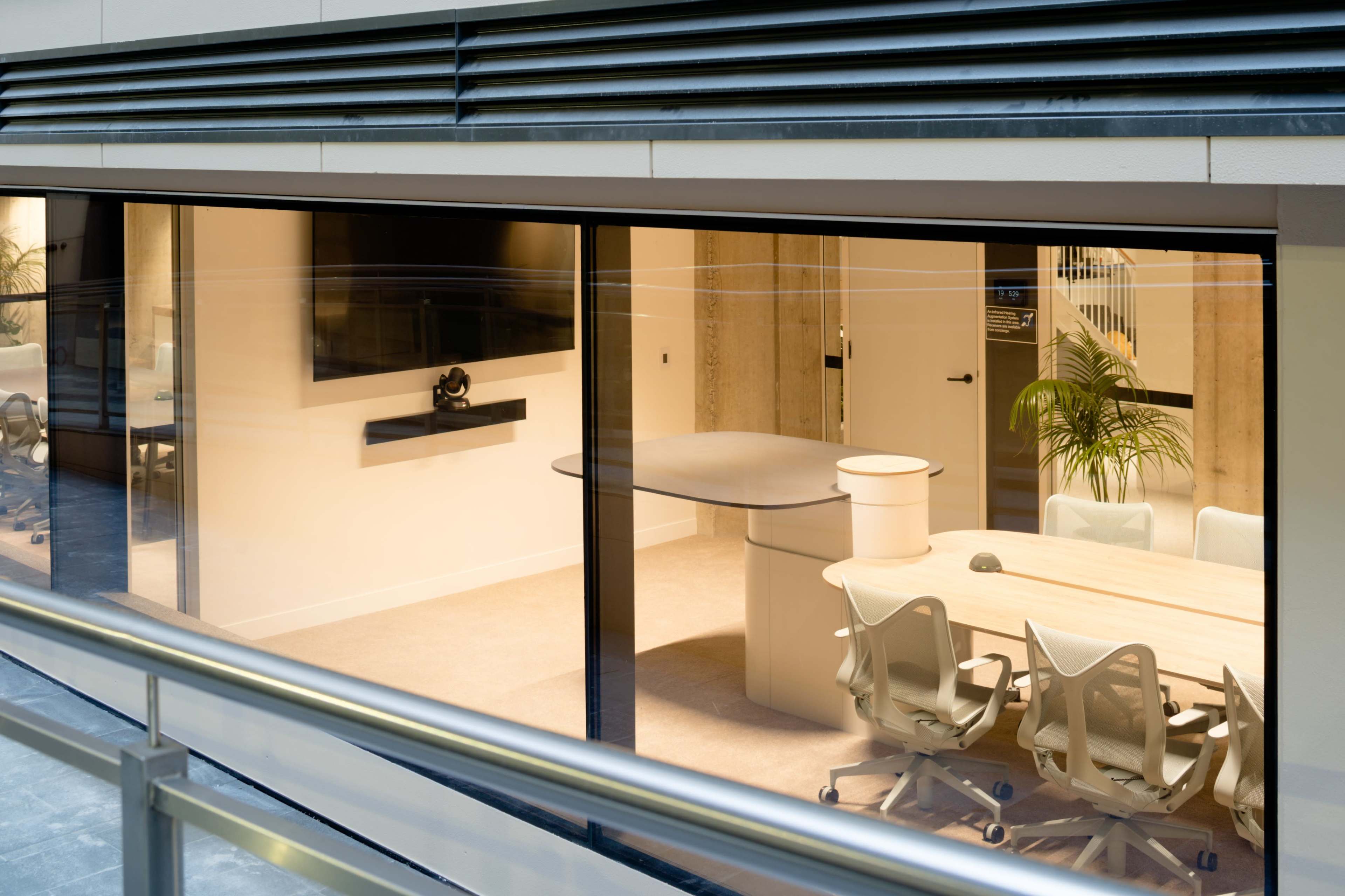 A modern conference room with a large table, several chairs, and a wall-mounted display, all enclosed in glass walls.