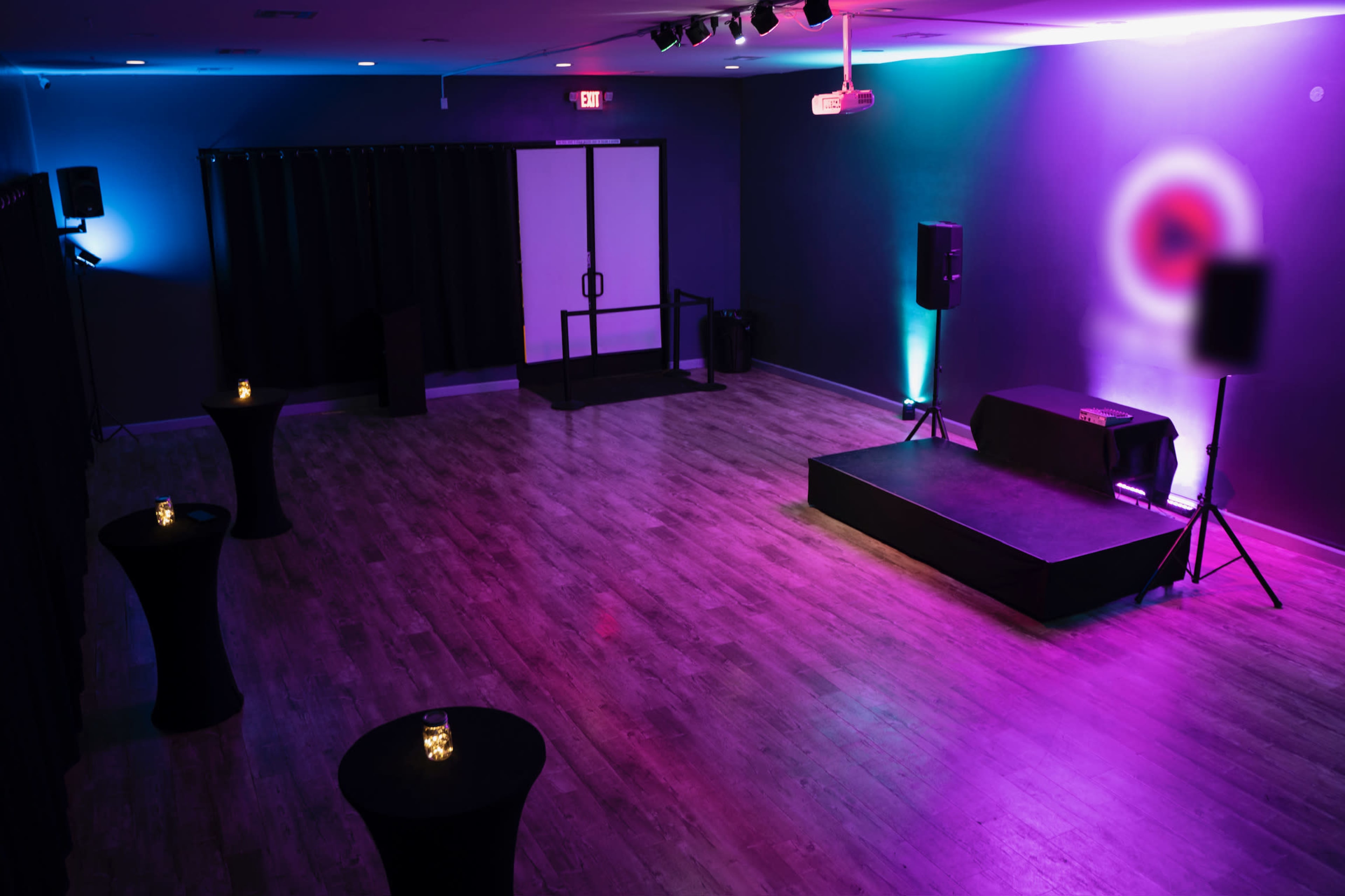 A room with a stage, spotlight, and several tables, illuminated by colorful lighting.