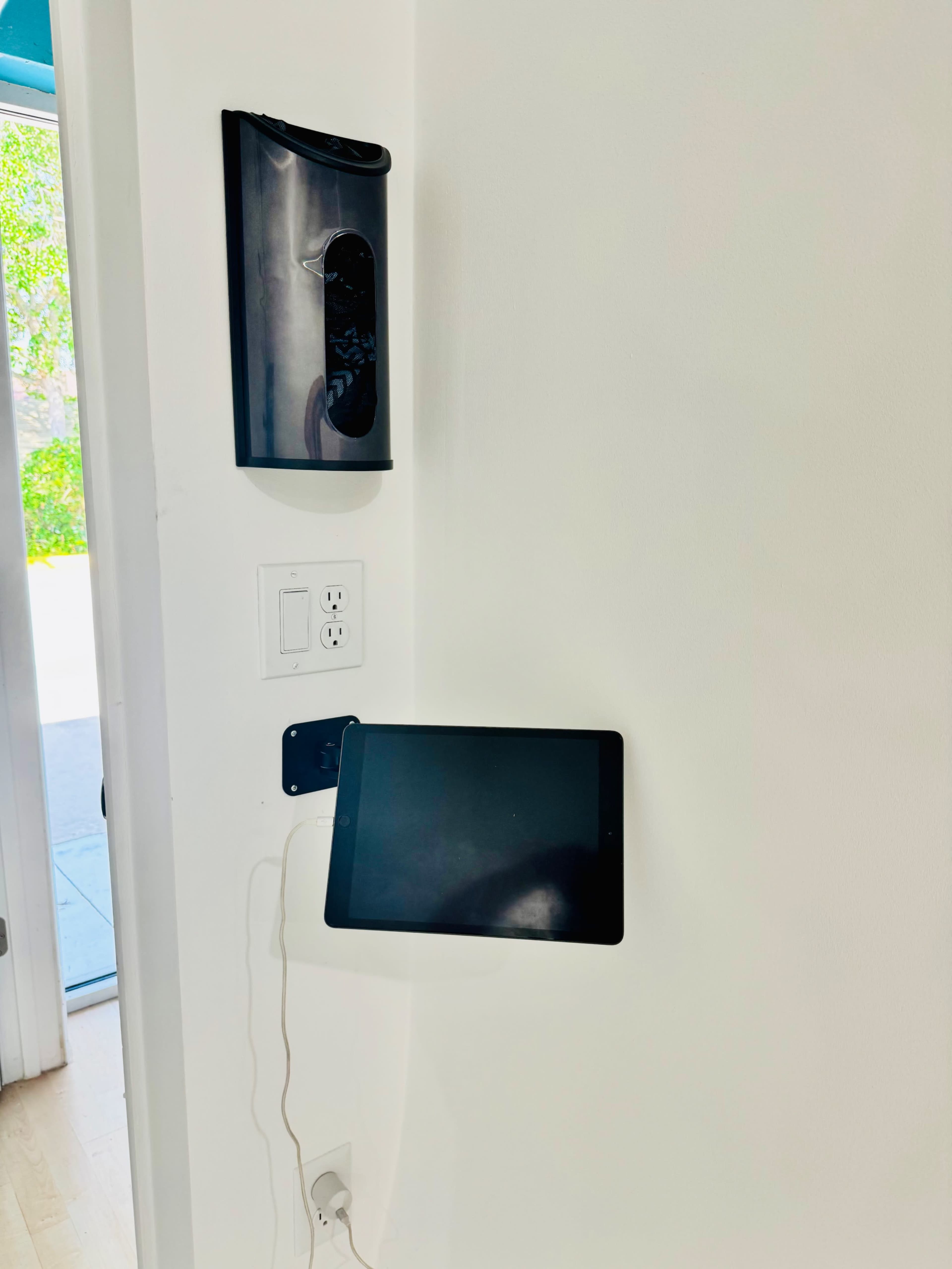 A black device is mounted on a white wall above a charging tablet that is attached to the wall, with a power outlet nearby.