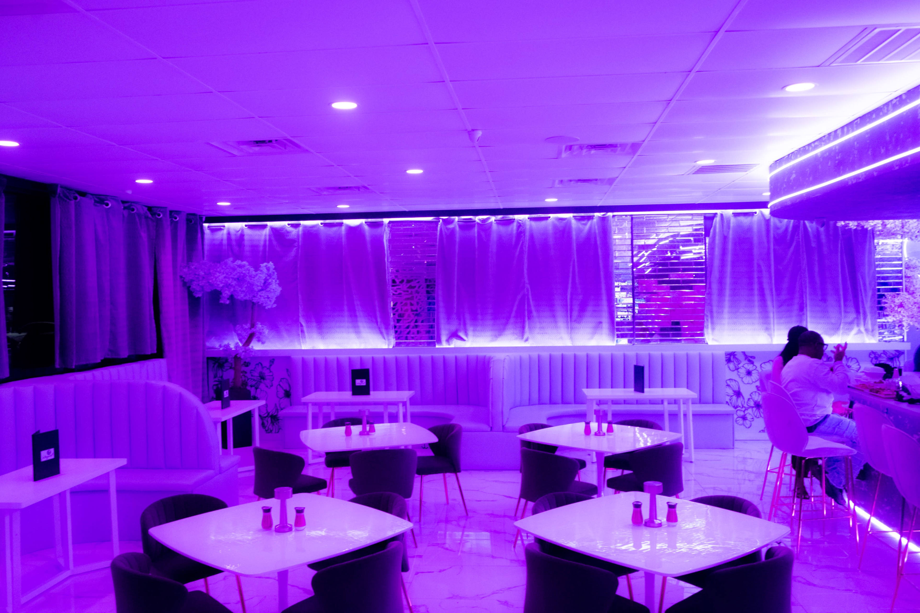 A modern café features purple lighting, white furniture, and multiple tables set for customers.