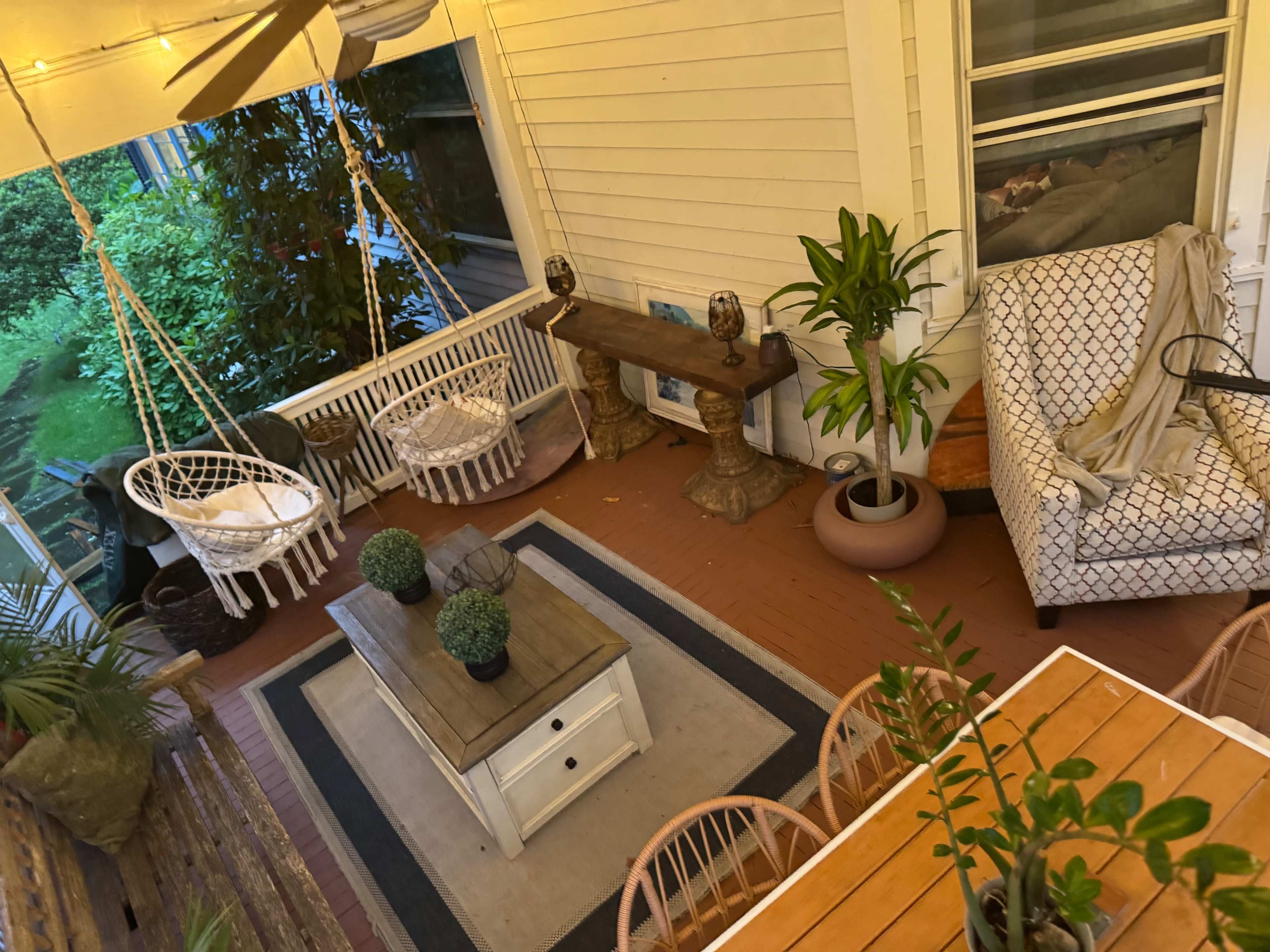 The image shows a cozy porch featuring a swing chair, a wooden table surrounded by chairs, potted plants, and a cushioned armchair.