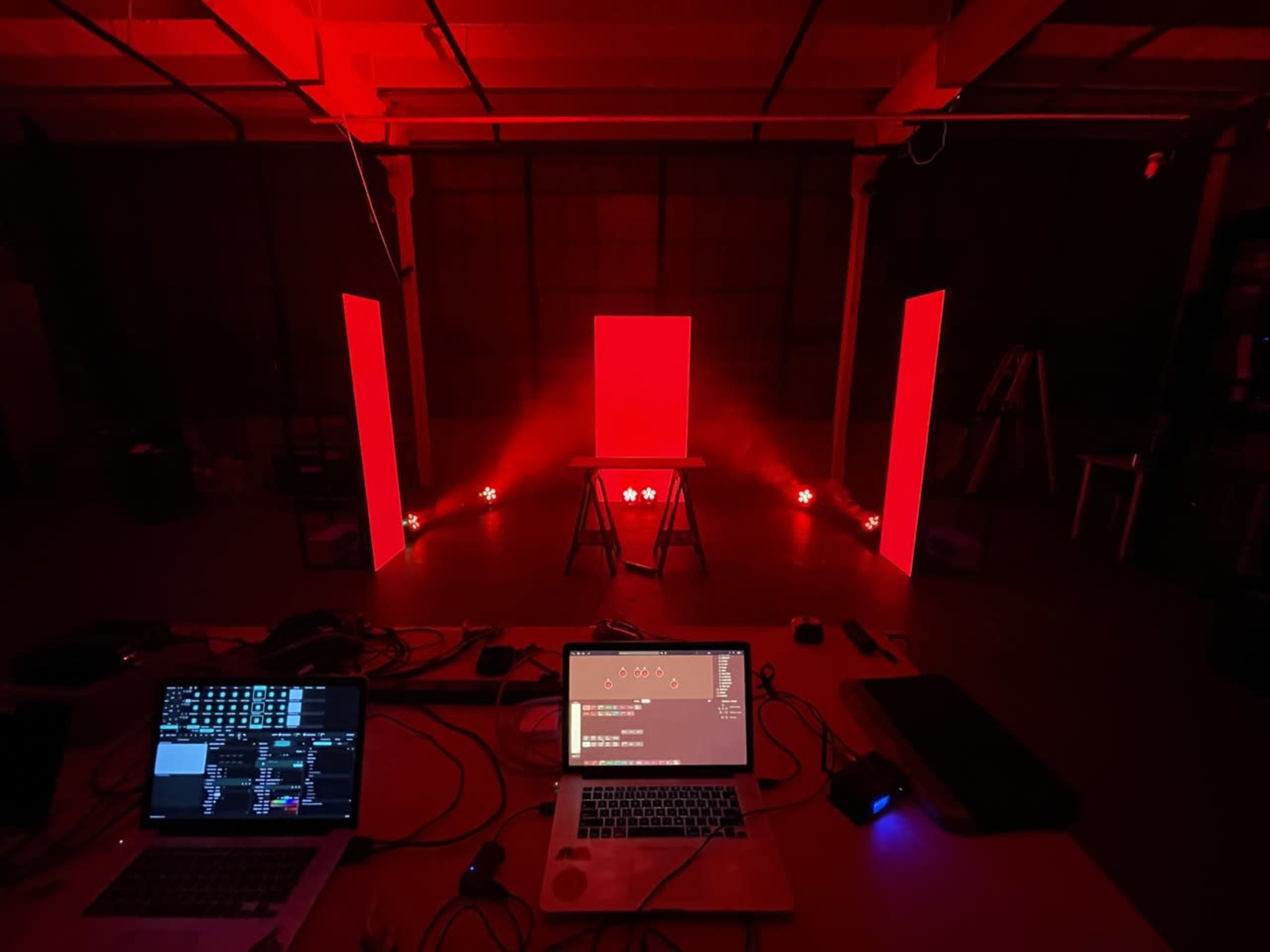 A dimly lit room features a central red light source on a pedestal, flanked by two red-lit panels, with a laptop and equipment scattered on a table in the foreground.
