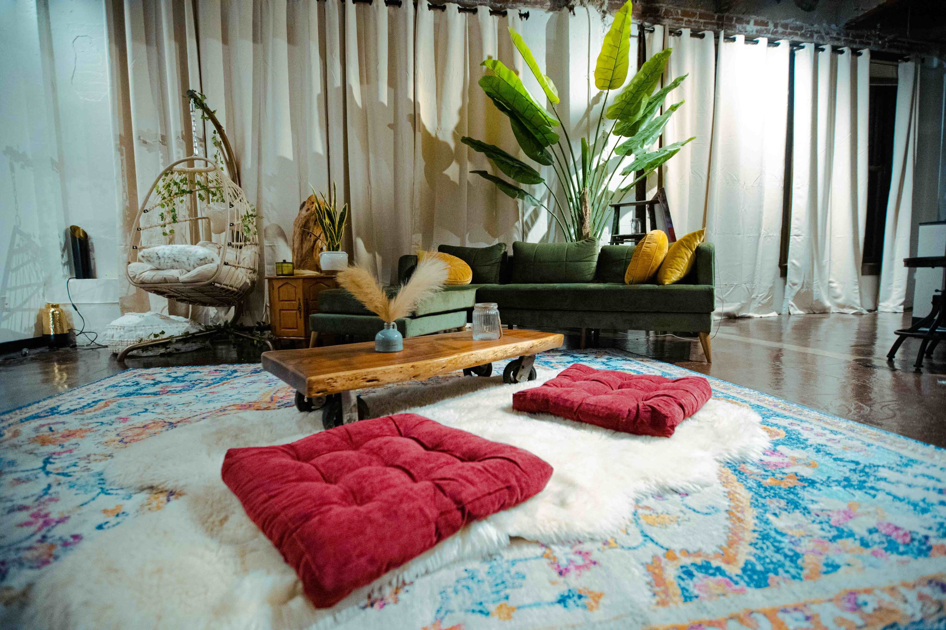 A cozy living room featuring a dark green sofa, a wooden coffee table, and several floor cushions arranged on a patterned rug, surrounded by plants and soft curtains.