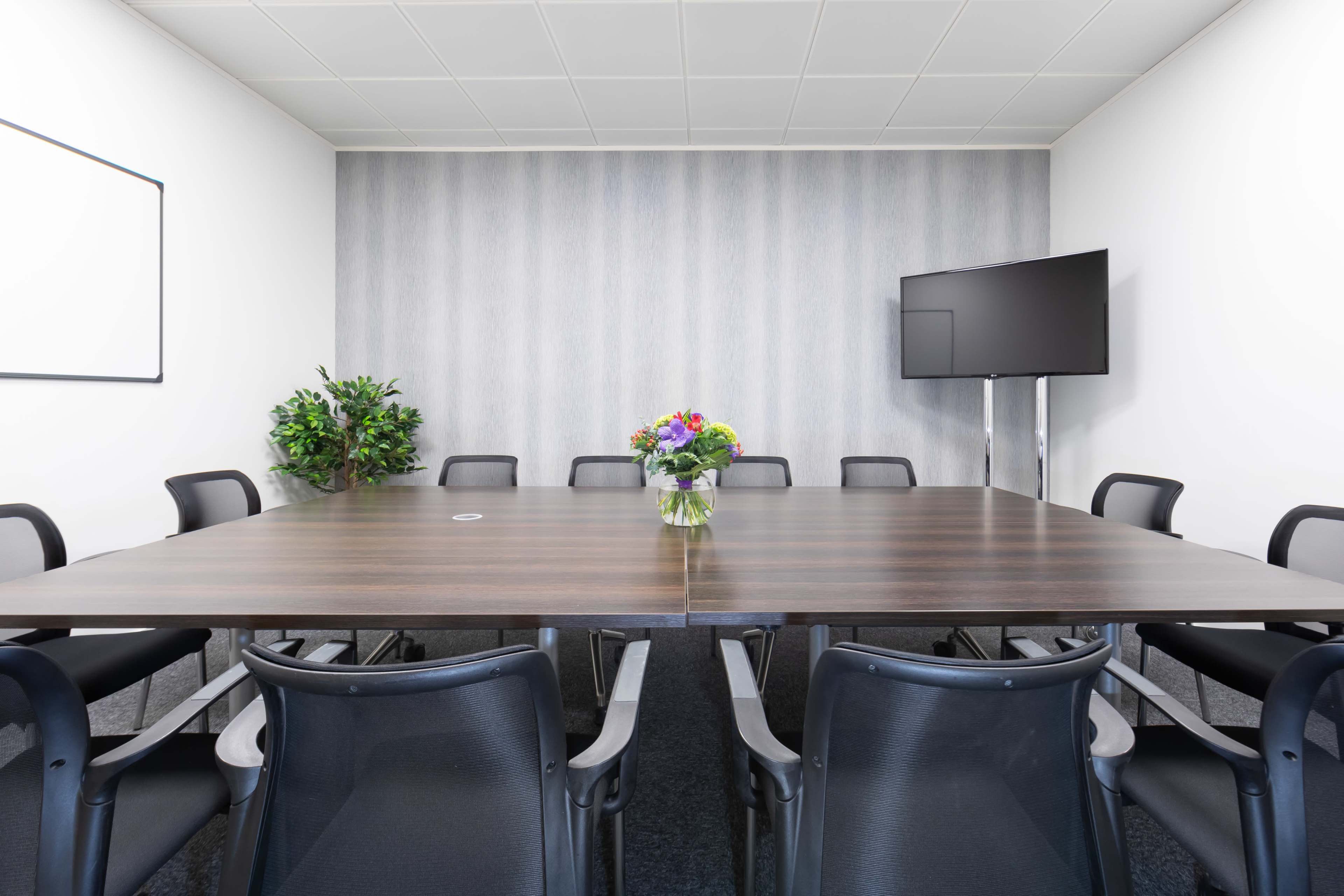 A conference room is set up with a large wooden table surrounded by black chairs, featuring a TV on a stand and a vase of flowers in the center.