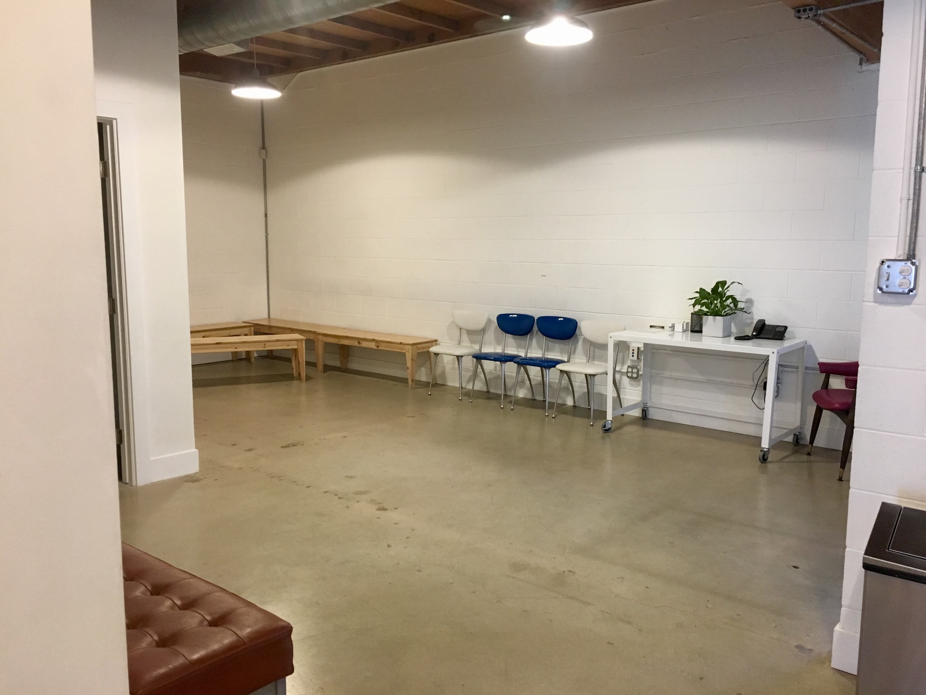 The image shows a spacious, minimalist indoor area with a wooden bench, several chairs, a table, and a potted plant against a white wall.