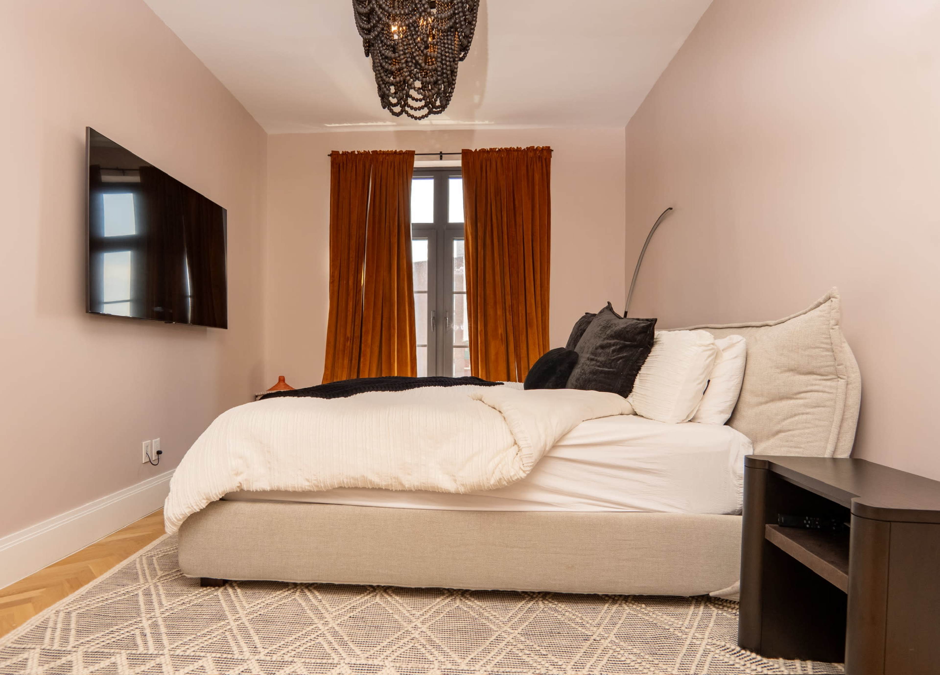 A minimalist bedroom features a large bed with white bedding, a mounted TV on the wall, and orange curtains.