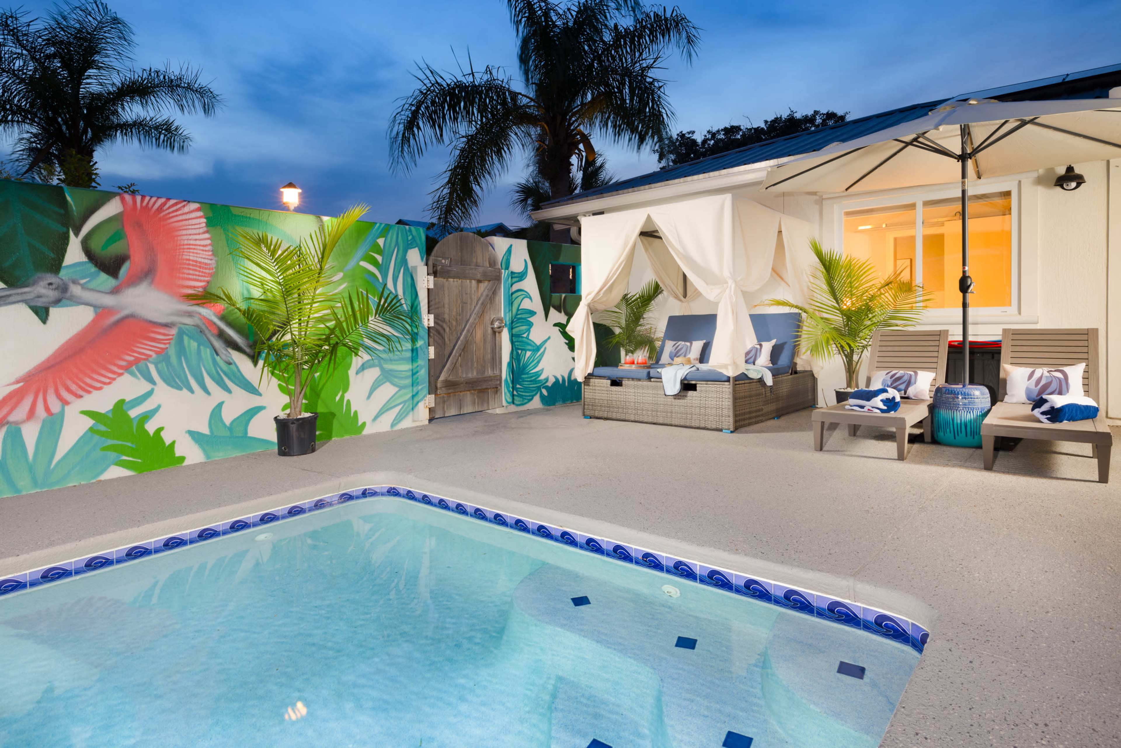 A pool area with a mural of a bird on the wall, lounge chairs with towels, and a shaded seating area beside the water.
