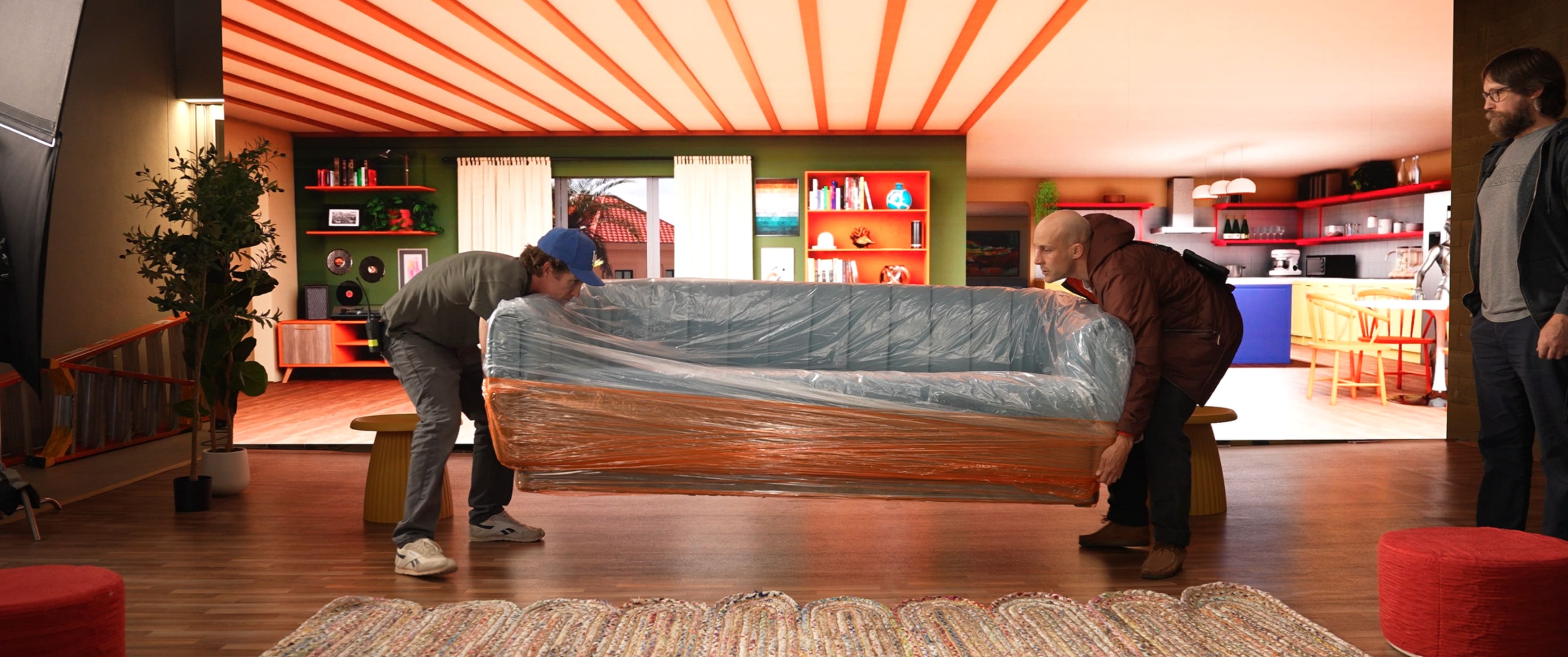 Two men are lifting a wrapped couch in a brightly colored interior room featuring shelves and furniture.