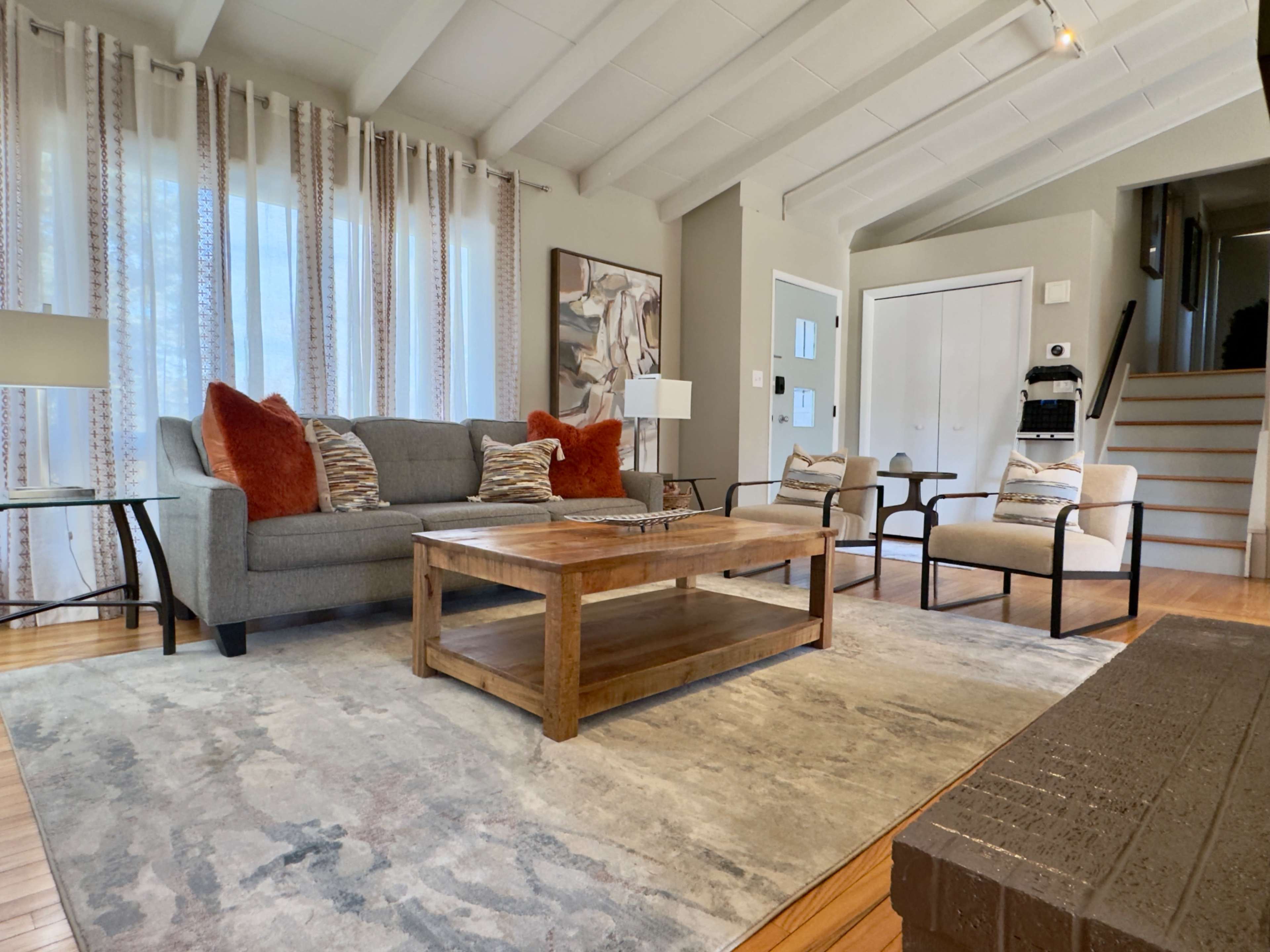 The living room features a gray sofa with decorative pillows, a wooden coffee table, and two armchairs, all arranged on a patterned rug under a ceiling with exposed beams.