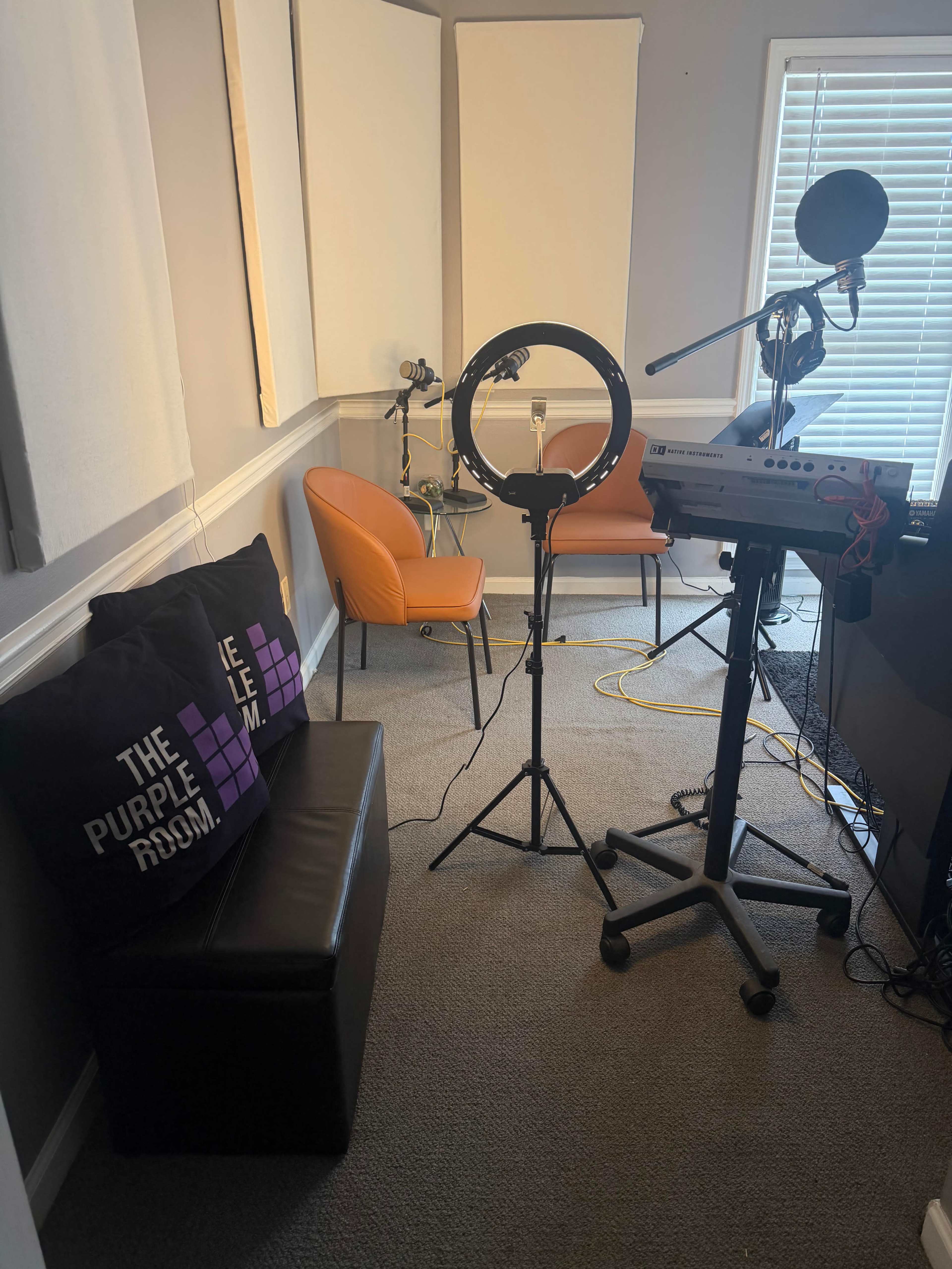 The image shows a small studio space featuring a keyboard, microphone, a ring light on a stand, two orange chairs, and a black bench with purple pillows against a gray wall.