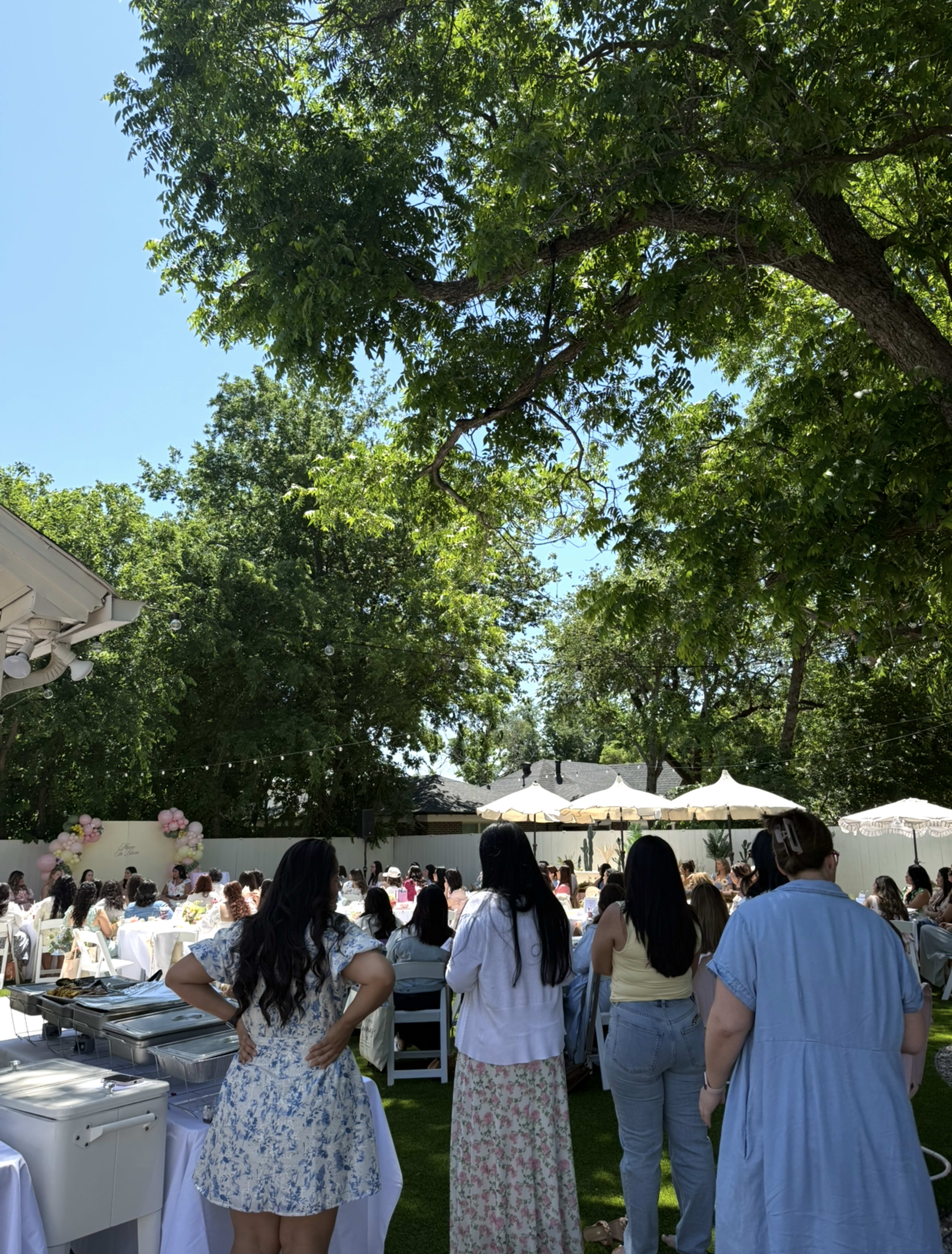 A large outdoor gathering is taking place under trees with attendees seated at tables, some standing and facing a speaker or presentation area.