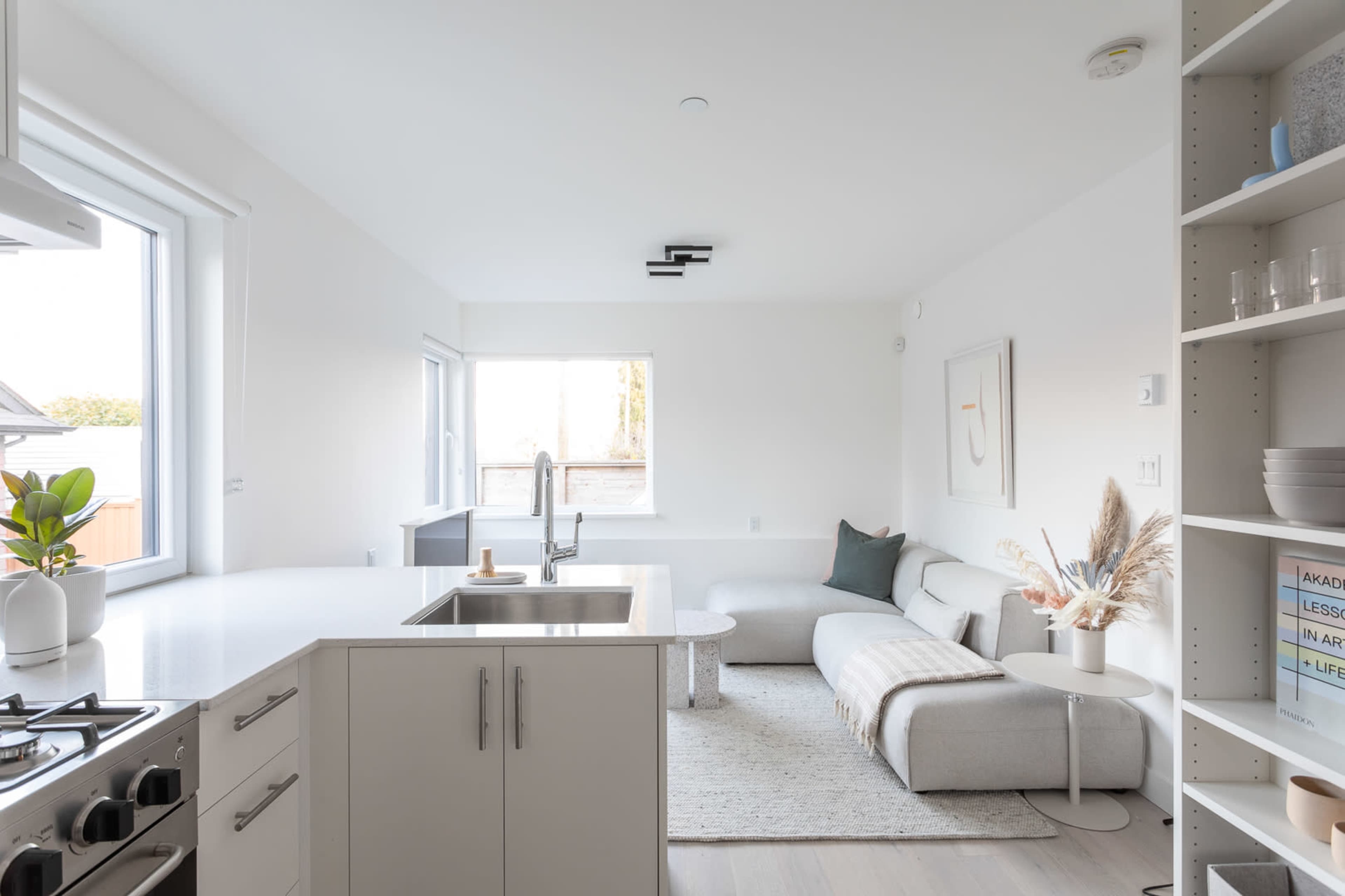 A bright, minimalist kitchen and living area features a gray sofa, a round coffee table, and large windows allowing natural light to enter.