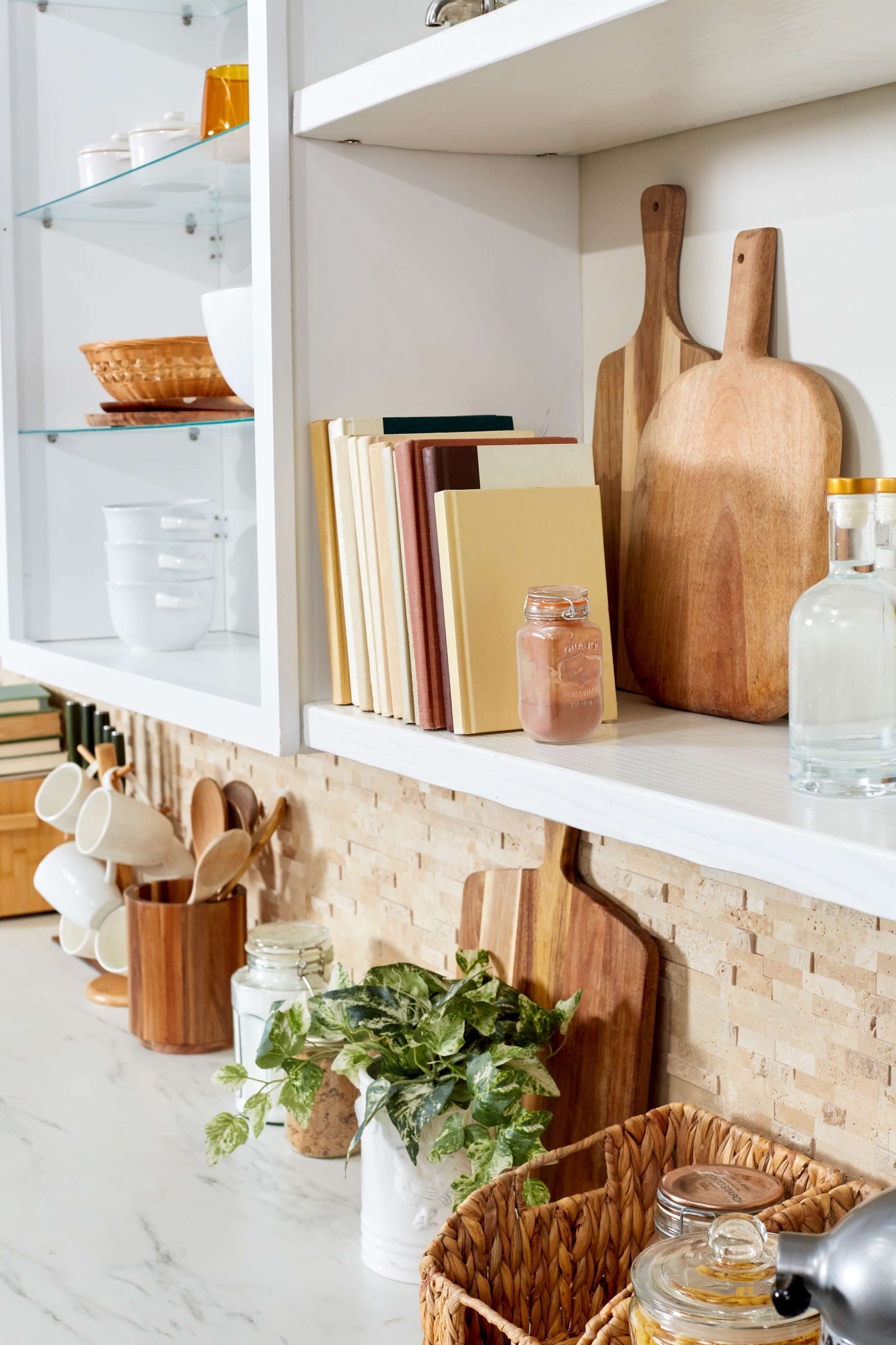 A neatly arranged kitchen shelf featuring wooden cutting boards, jars, books, and glassware against a tiled backdrop.