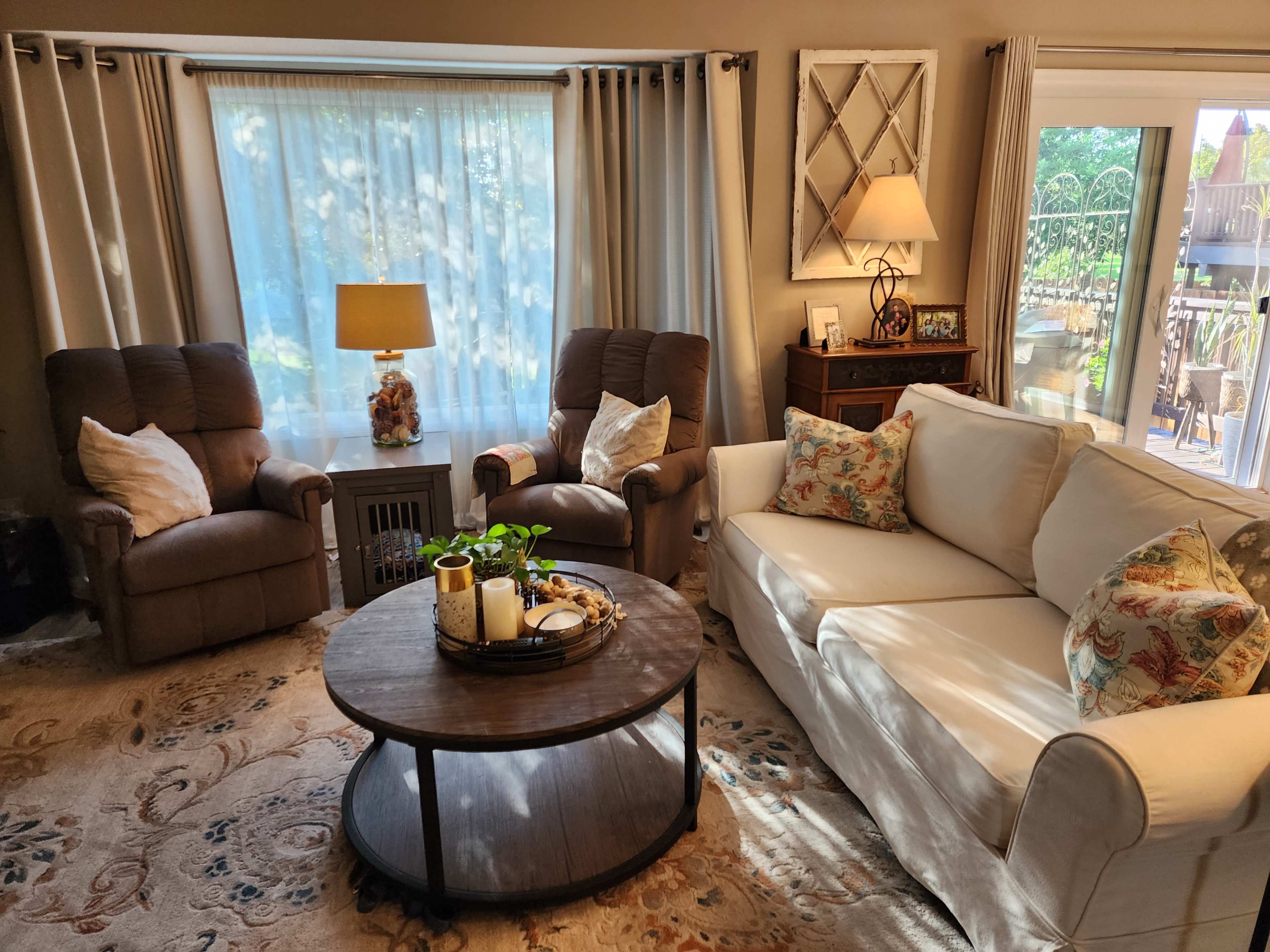 The image shows a cozy living room with two recliners, a light-colored sofa, a round coffee table adorned with decorative items, and large windows letting in natural light.