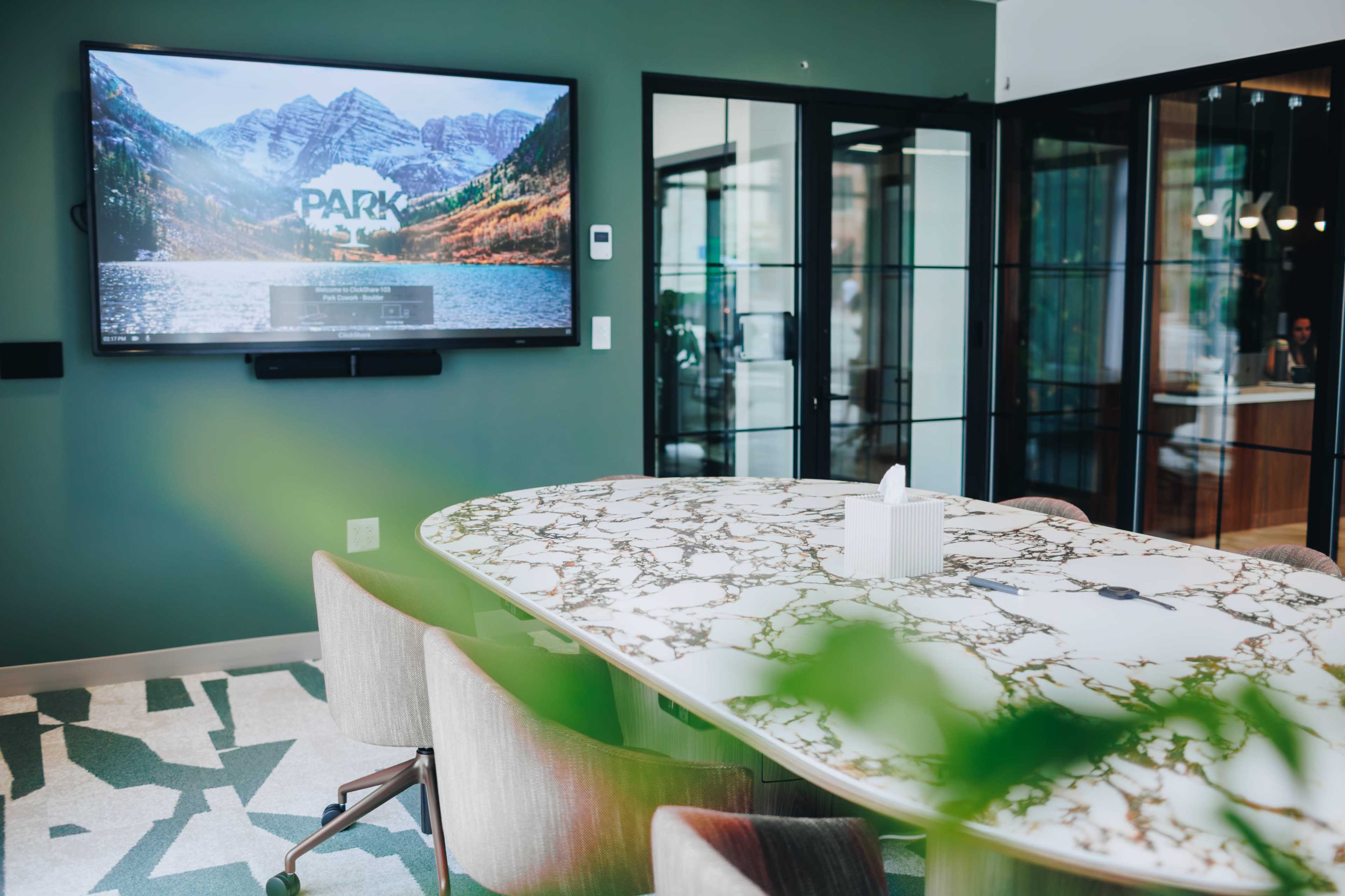 A conference room features a large marble table surrounded by upholstered chairs, with a screen displaying a scenic mountain landscape on the wall.