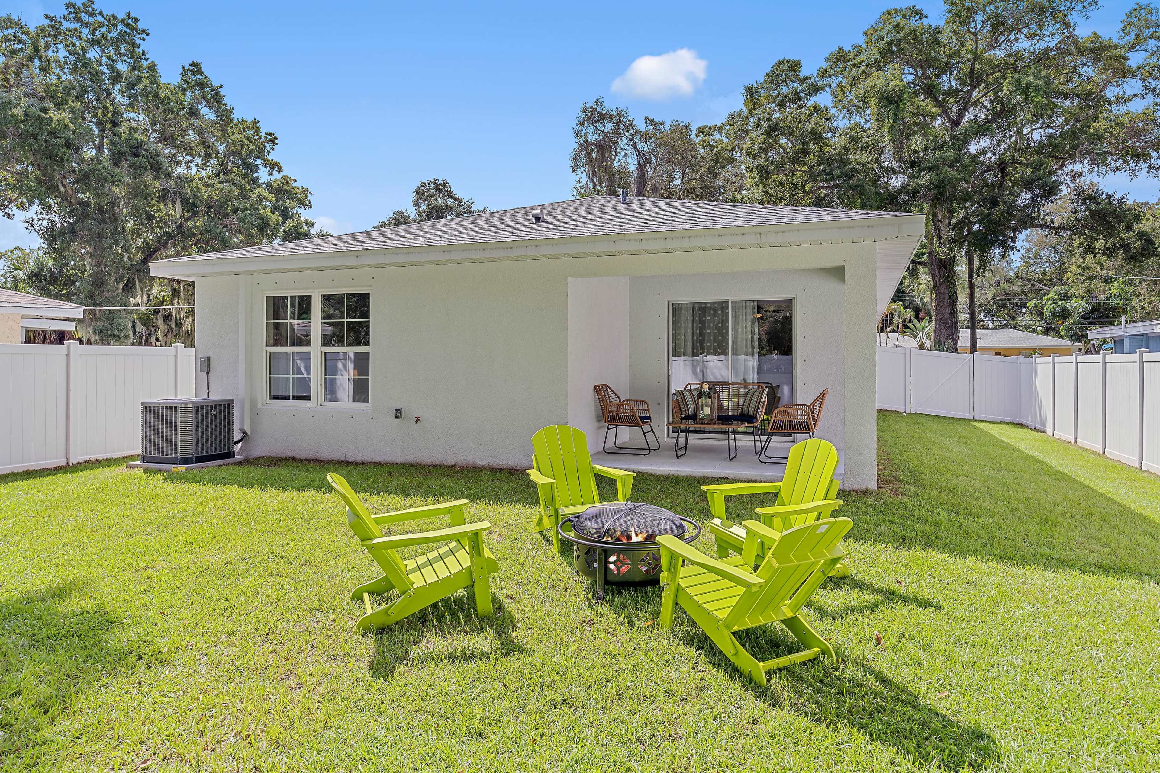 A back yard with a fire pit surrounded by four green chairs, a small table, and a view of a house with large windows.