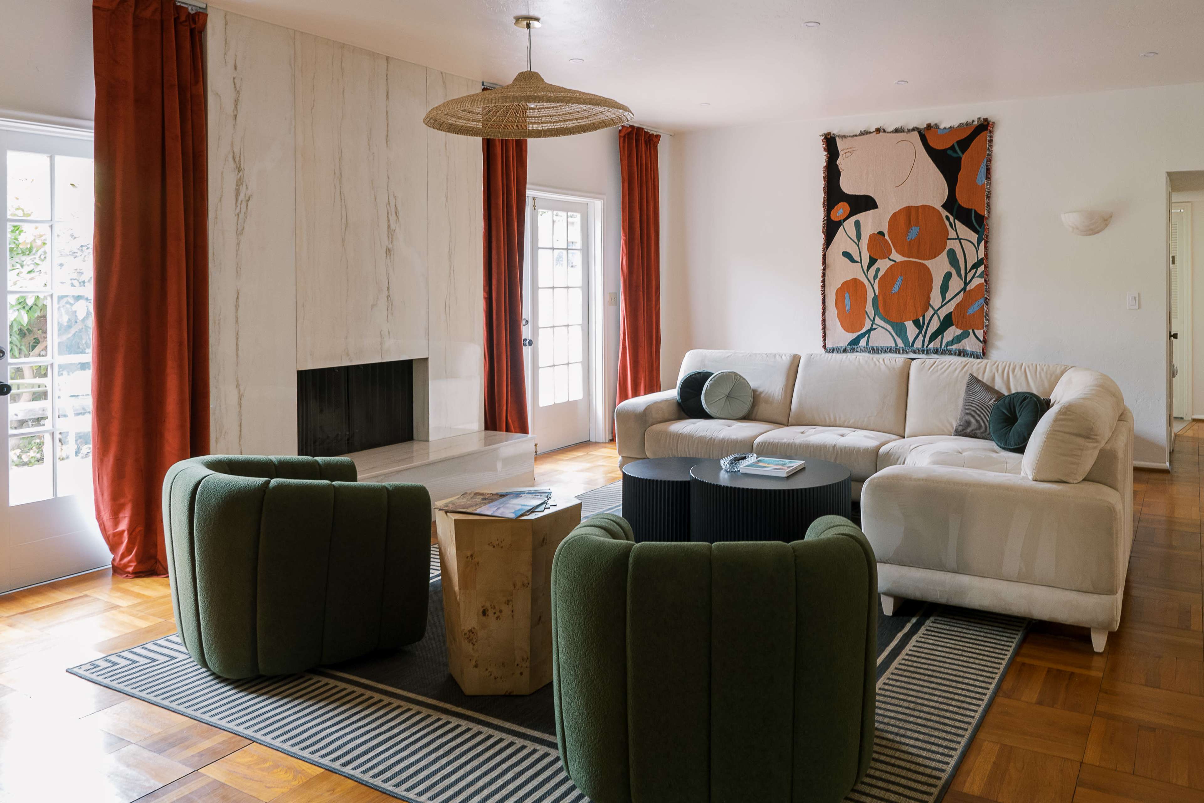 A modern living room features a white sectional sofa, green accent chairs, a round black coffee table, and a marble fireplace, with large windows dressed in red curtains.