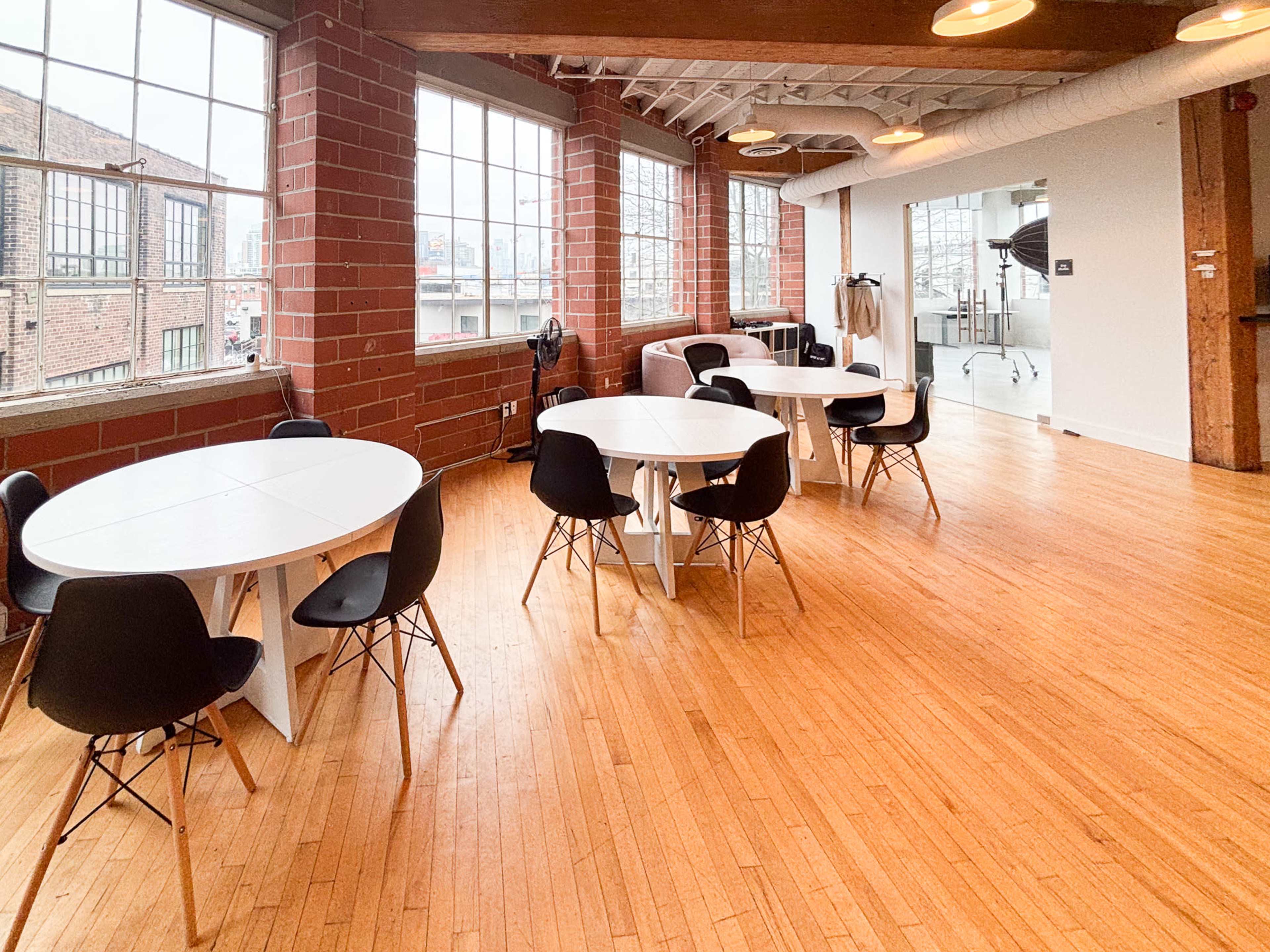 The image shows a spacious room with wooden flooring, featuring round tables with black chairs, large windows, and a modern design.