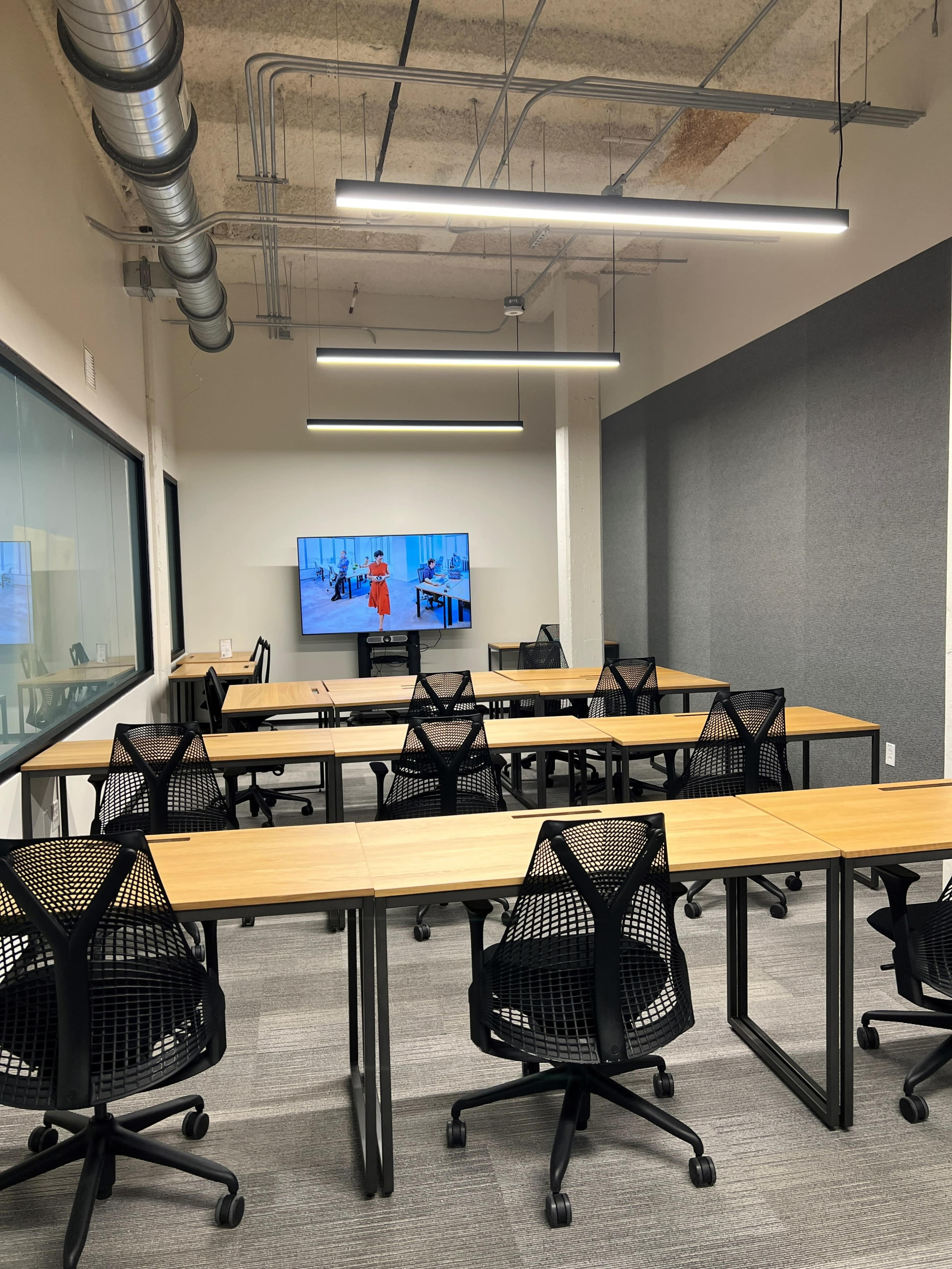 A modern conference room features multiple tables and chairs arranged with a large screen displaying a presentation on one wall.